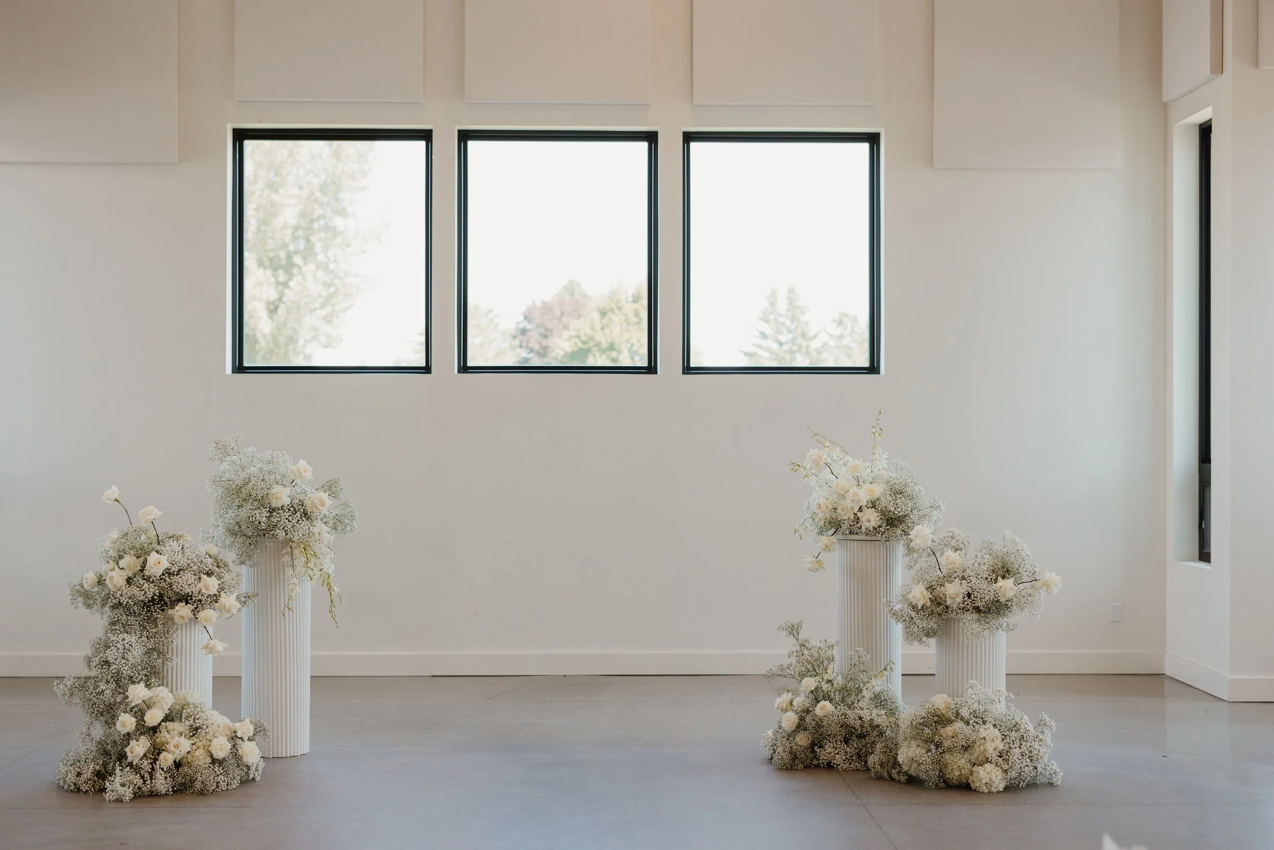 Elegant indoor setting with three tall white floral arrangements on white pedestal vases, decorated with white flowers and baby's breath, positioned on a smooth floor against a plain white wall with three large windows showing outdoors.