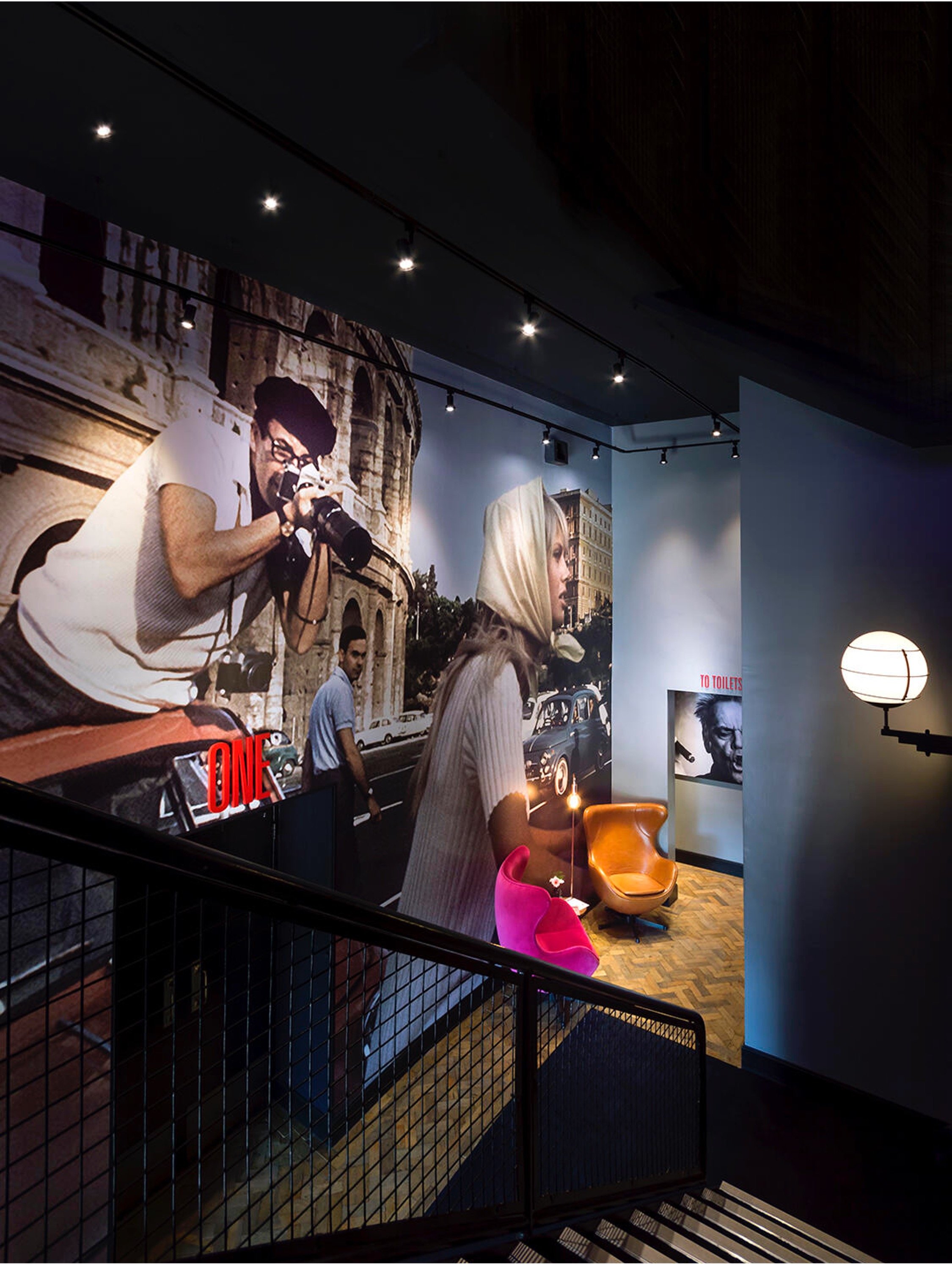 Interior of a museum or gallery with a large wall mural featuring vintage street scene and portraits, including a woman with a white headscarf and a photographer taking pictures, with colorful chairs and a lamp in the foreground.