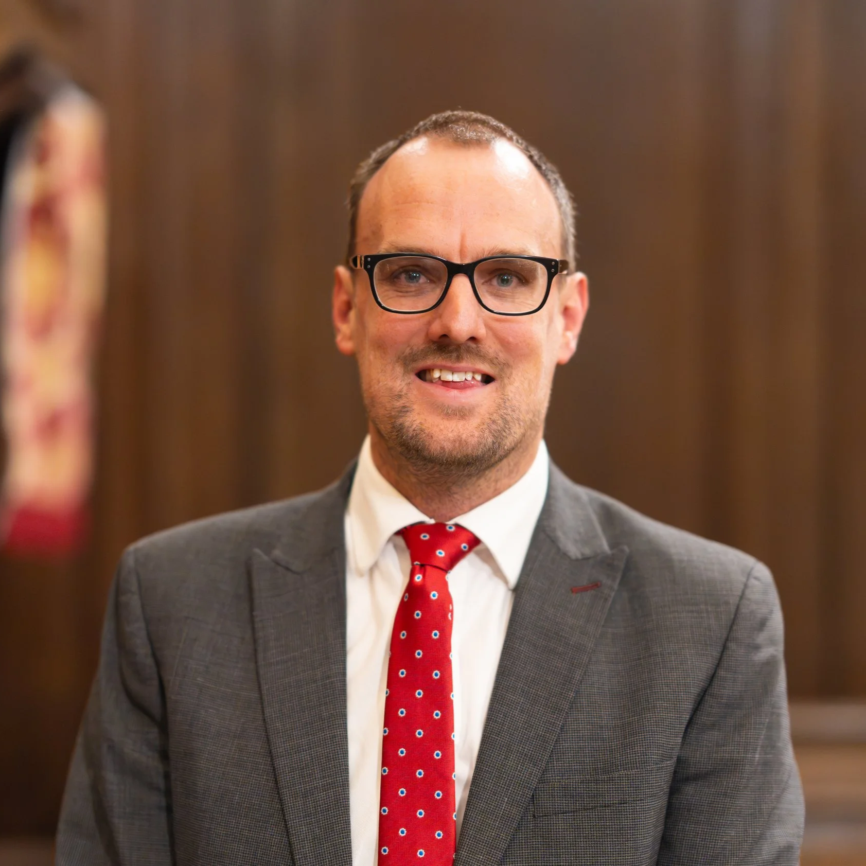 A man in glasses with short dark hair, wearing a gray suit, white shirt, and red tie with blue and white polka dots, smiling in front of a brown wooden wall.