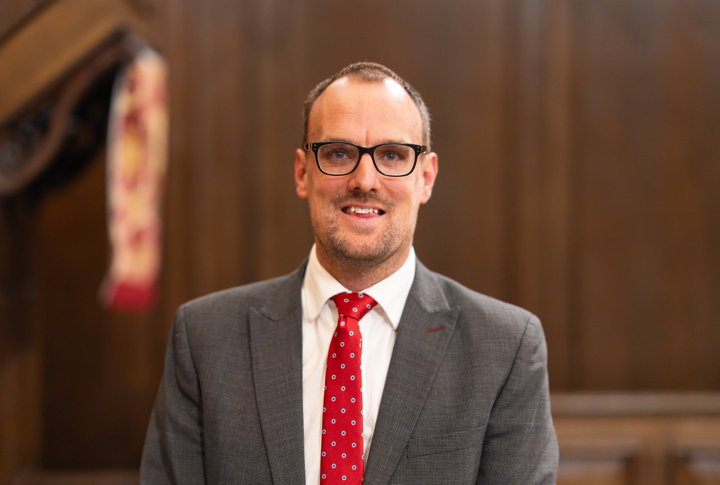 A man wearing glasses, a gray suit, a white shirt, and a red tie with blue dots, standing in a room with wooden paneling in the background.