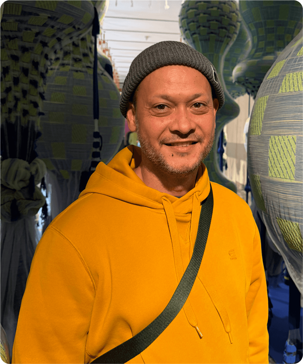 Ein Mann trägt eine graue Mütze und eine gelbe Kapuzenjacke, steht in einer Halle mit Marinetypen, vermutlich Yokohama, im Hintergrund.