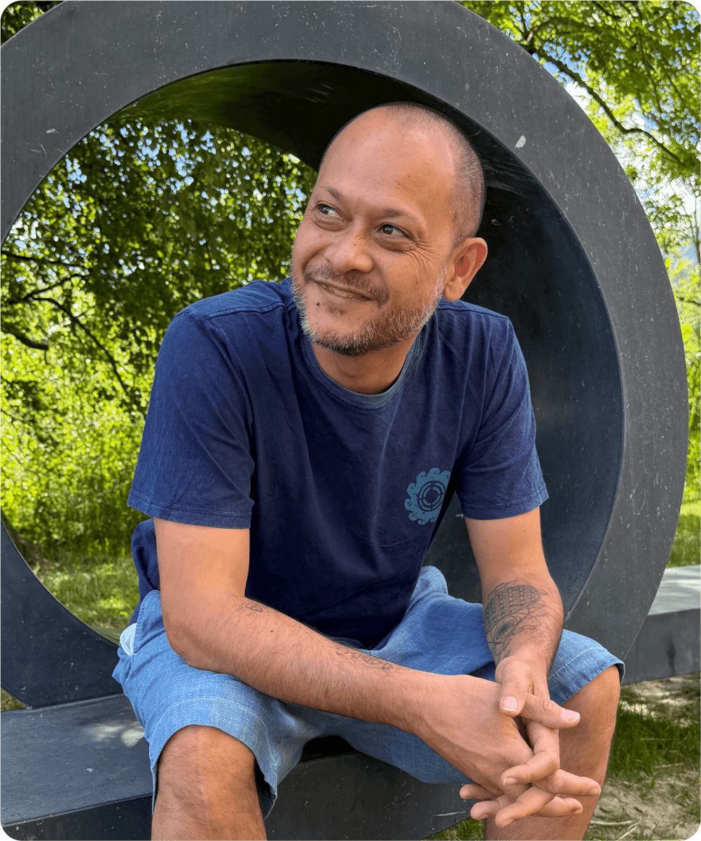 Ein Mann mit kurzhaar, Bart und Tattoo sitzend vor einer großen schwarzen Skulptur im Grünen, bei Sonnenschein.