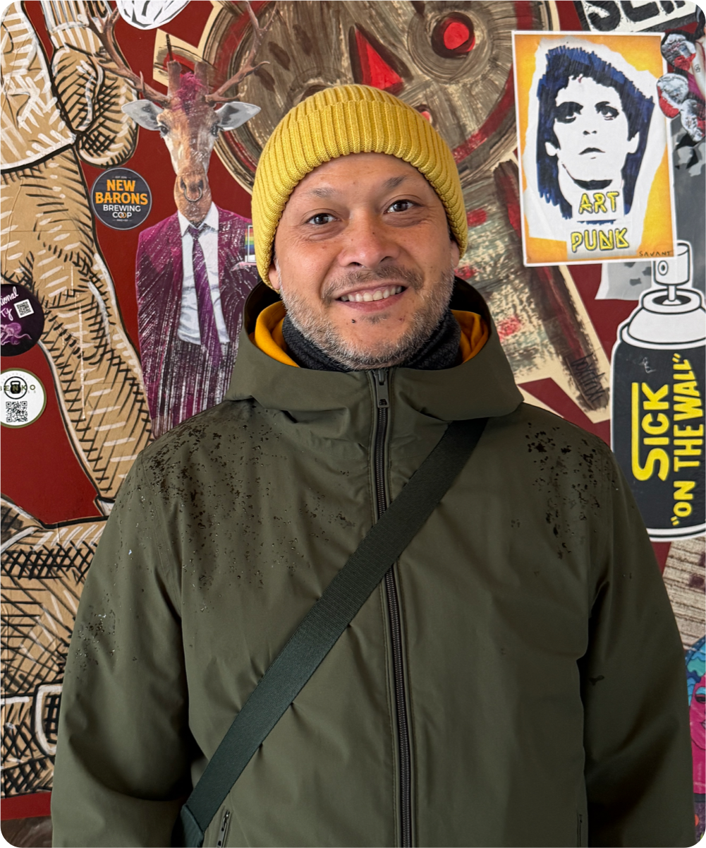 Ein Mann trägt eine gelbe Mütze, eine grüne Jacke und steht vor einer lebhaften Wand mit verschiedenen Graffiti und Street Art, darunter ein Hirsch mit Anzug, ein Bild im Punk-Stil und ein Spraydosen-Design.