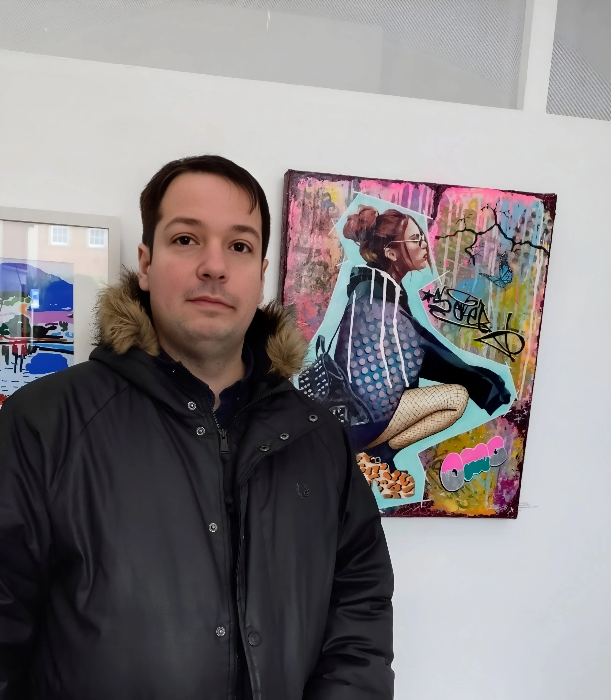 A man in a black winter jacket standing in front of a colorful graffiti-style artwork of a woman with glasses, hair in a bun, and wearing fishnet stockings, seated with one leg crossed.