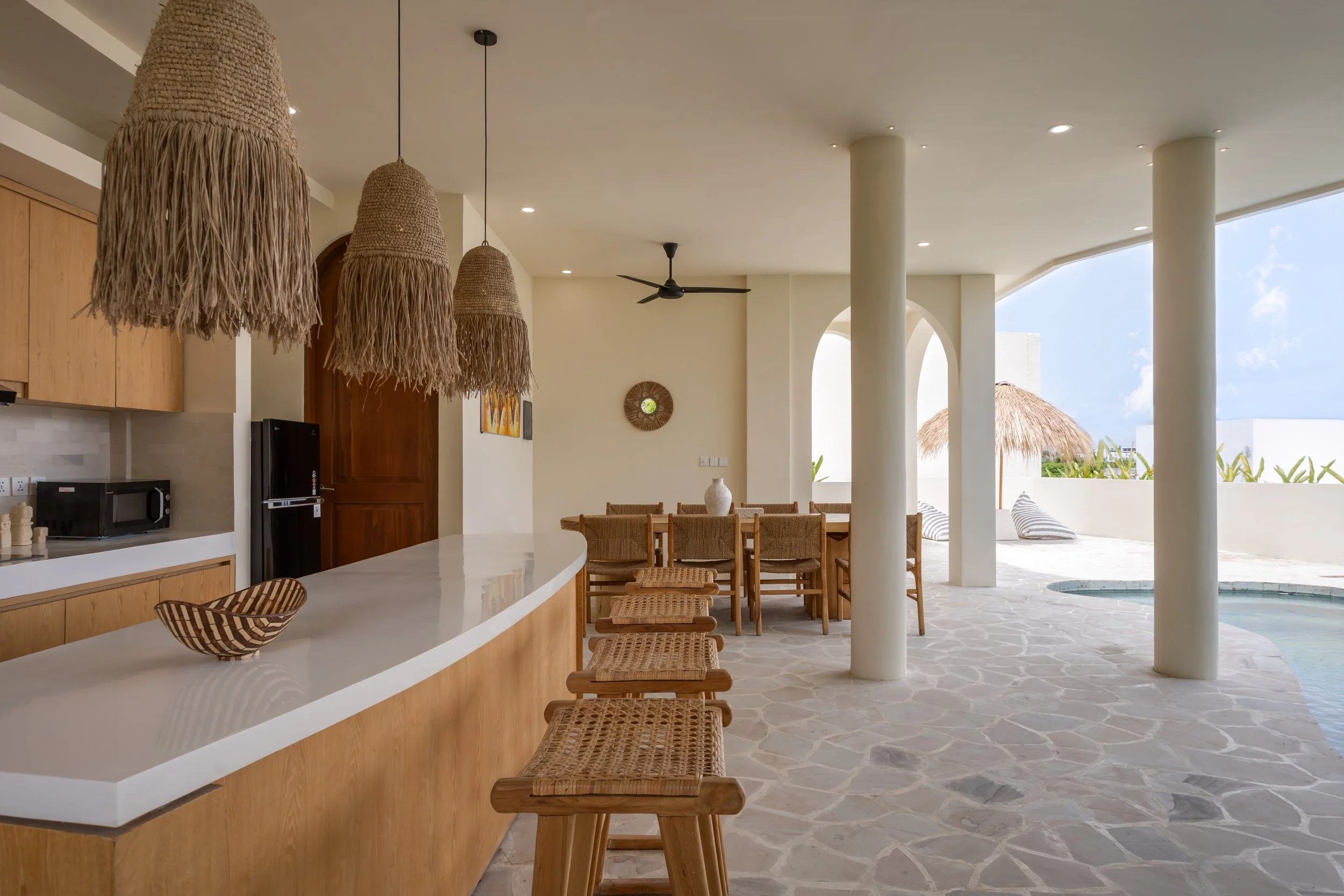 Open-air kitchen and dining area with a curved white countertop, wicker barstools, dining table with chairs, hanging straw pendant lights, and an outdoor lounge with striped pillows near a small pool, with a view of thatched umbrellas and blue sky.