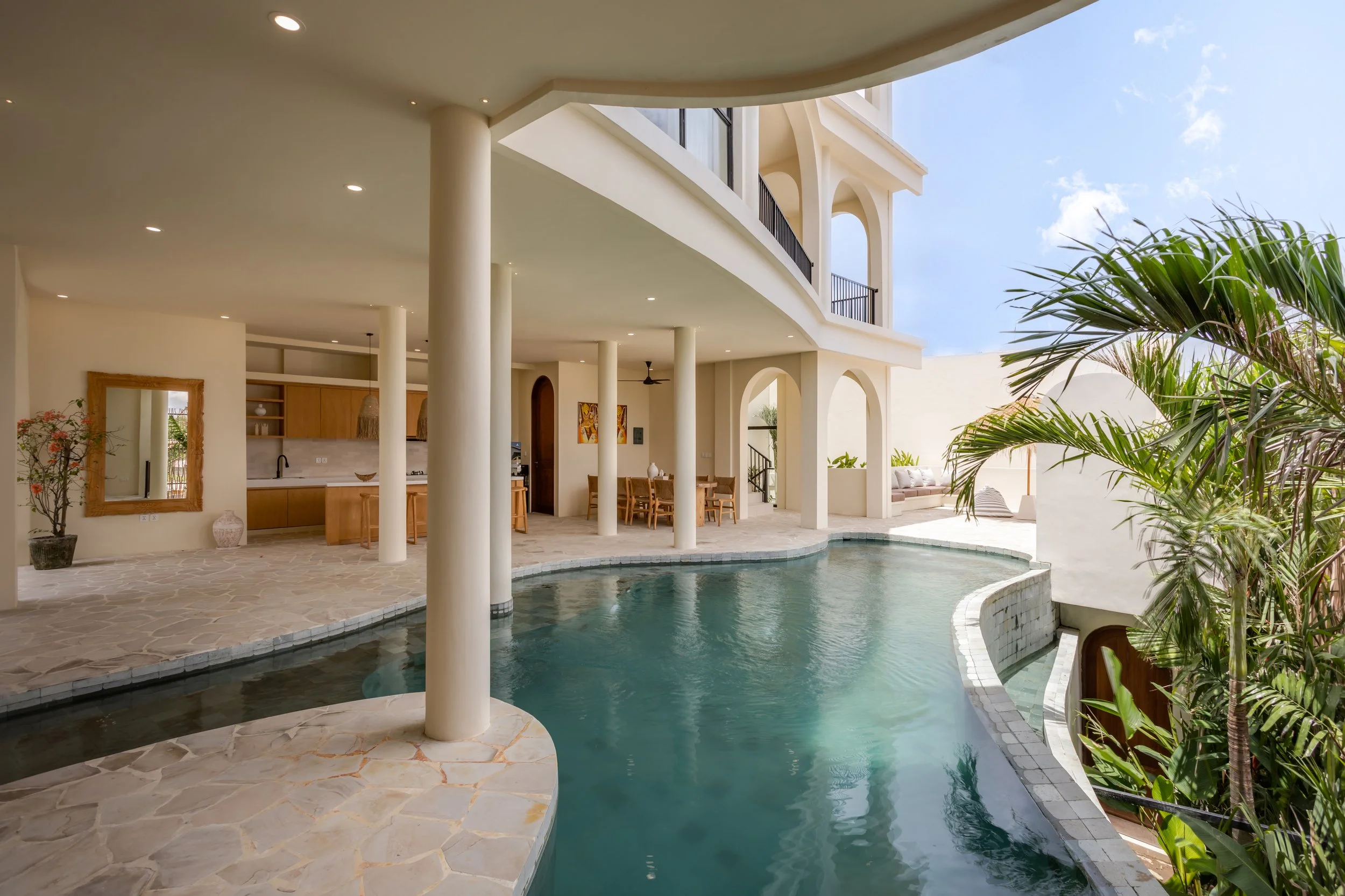 Indoor-outdoor pool area of a modern house with lush green plants, a curved swimming pool, and a patio with furniture, under a clear blue sky.
