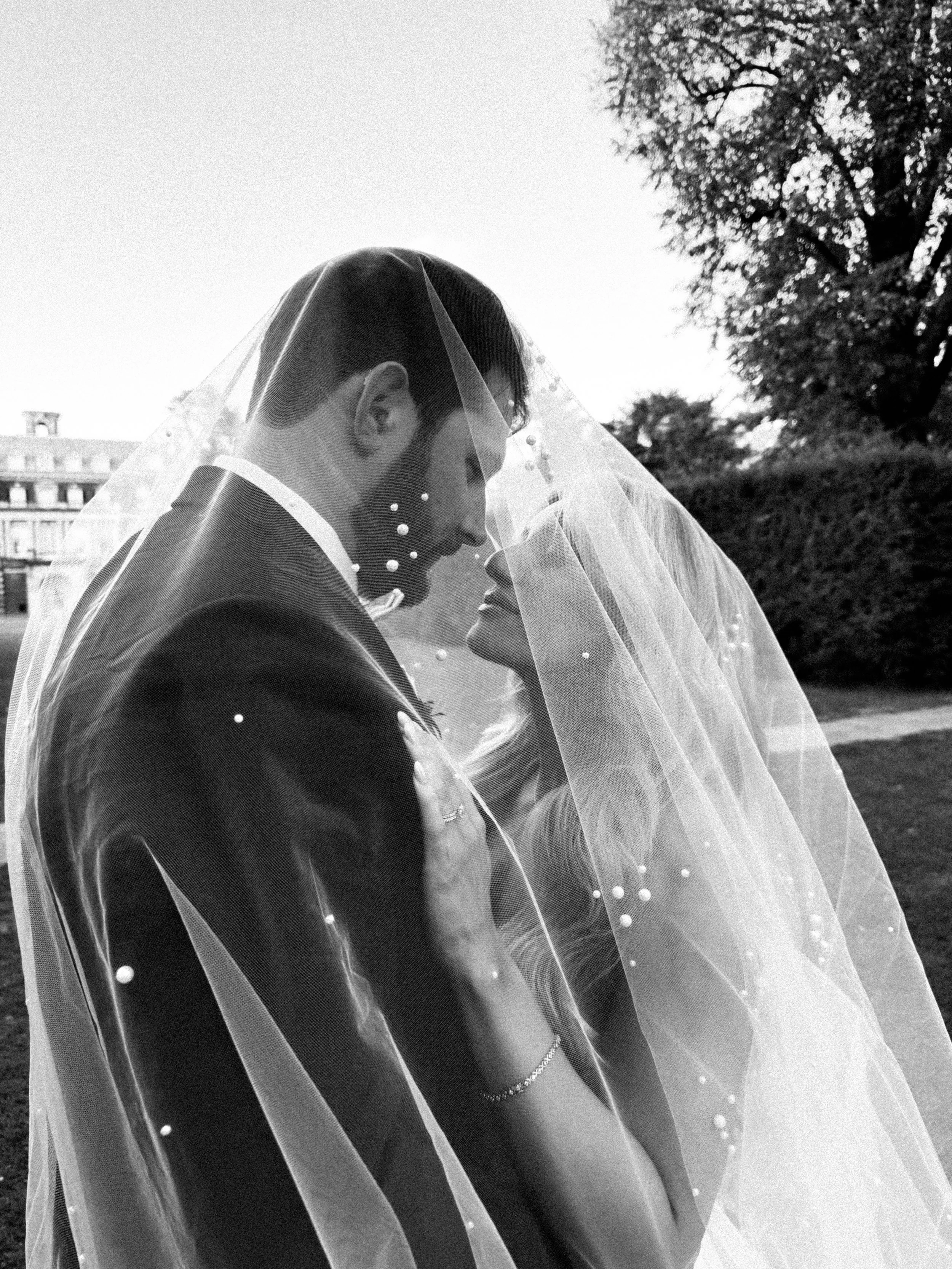 A black and white photo of a bride and groom under a veil, sharing a romantic moment outdoors with trees and a building in the background.