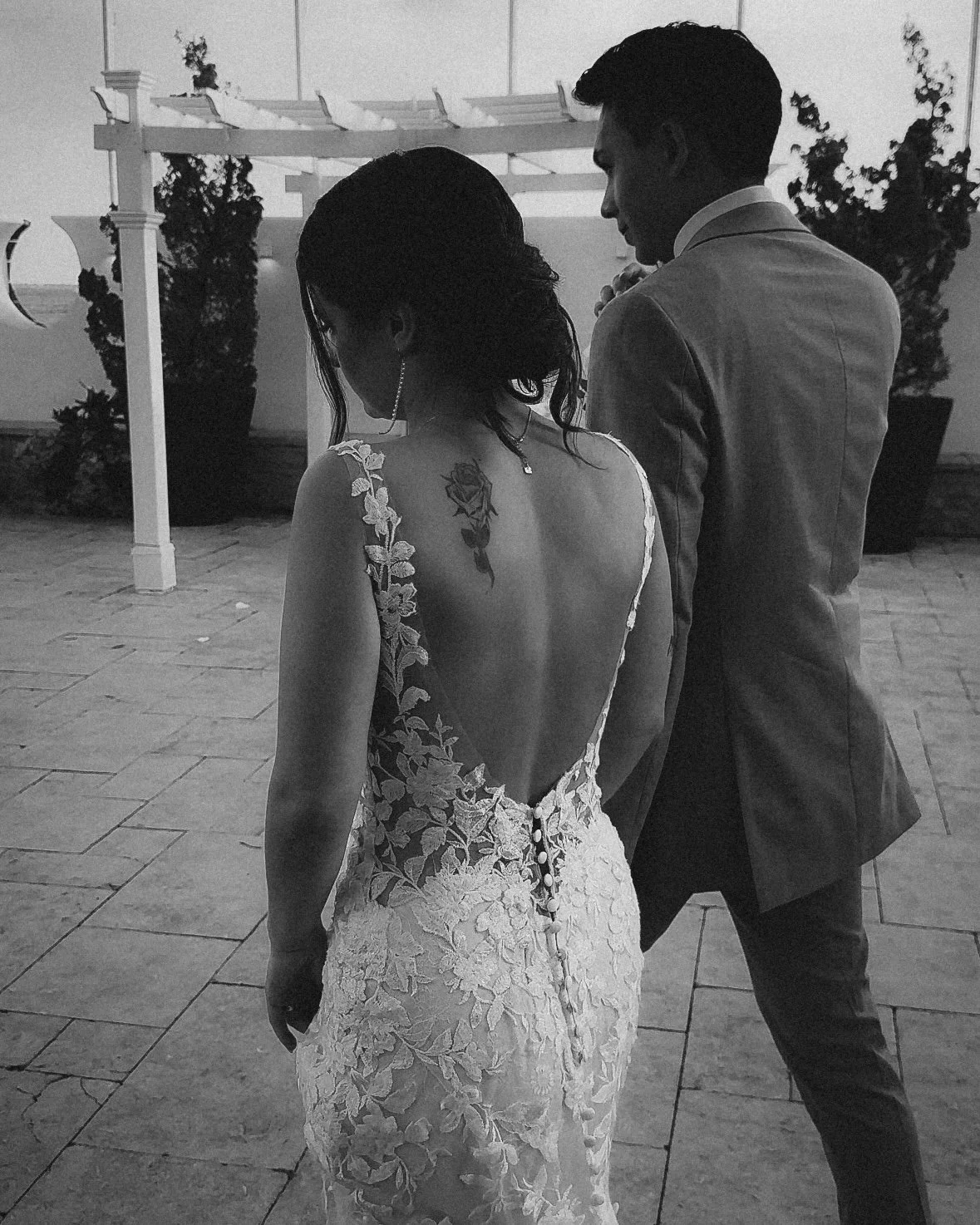 A bride and groom stand together outside during sunset, with their backs facing the camera. The bride is wearing a lace wedding dress with a low back and a tattoo on her shoulder. The groom is in a suit.