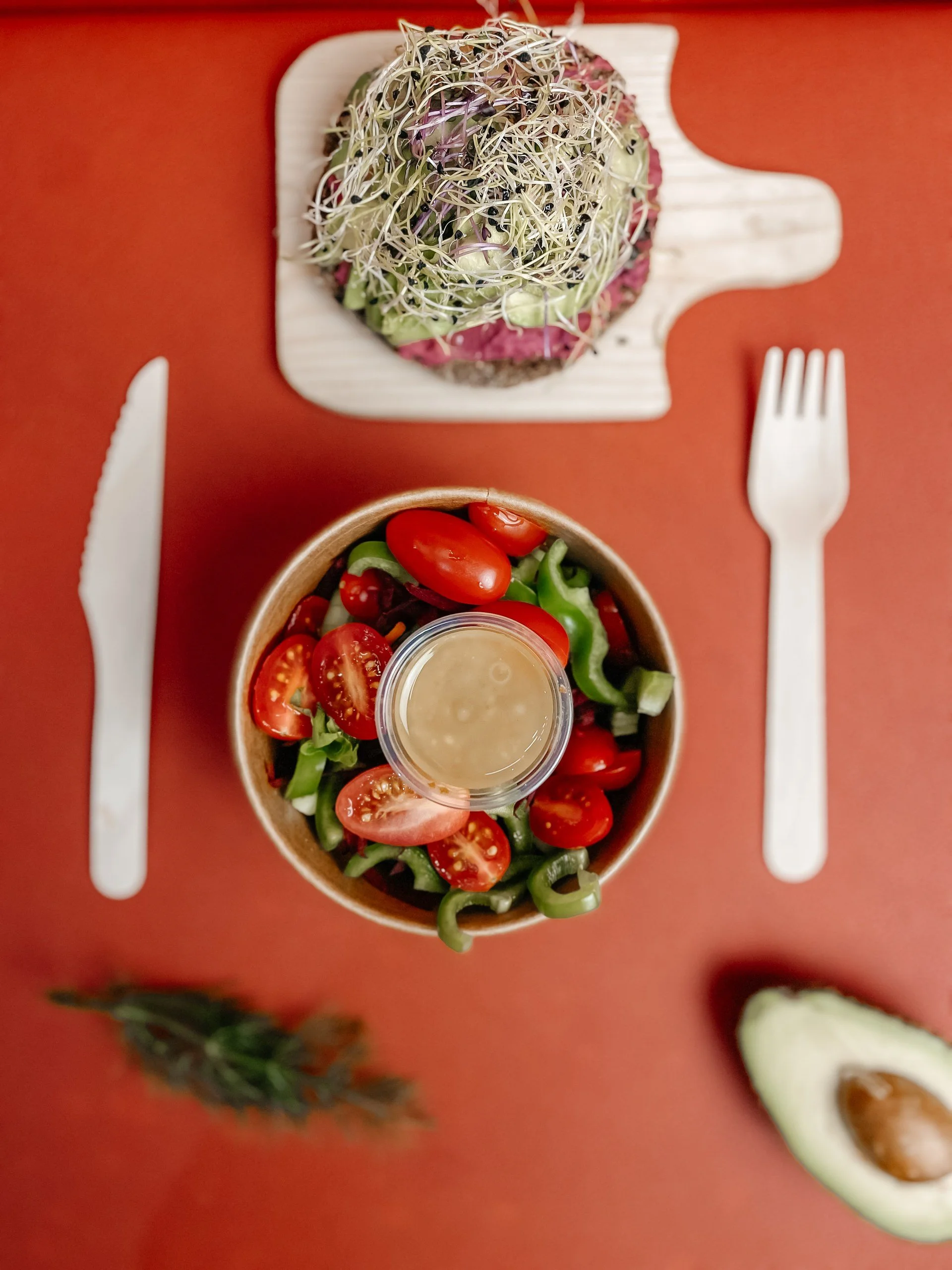 A salad bowl with cherry tomatoes, green bell peppers, and a small container of salad dressing on a red table, with a side of sliced avocado, a sprig of rosemary, a knife, and a fork.