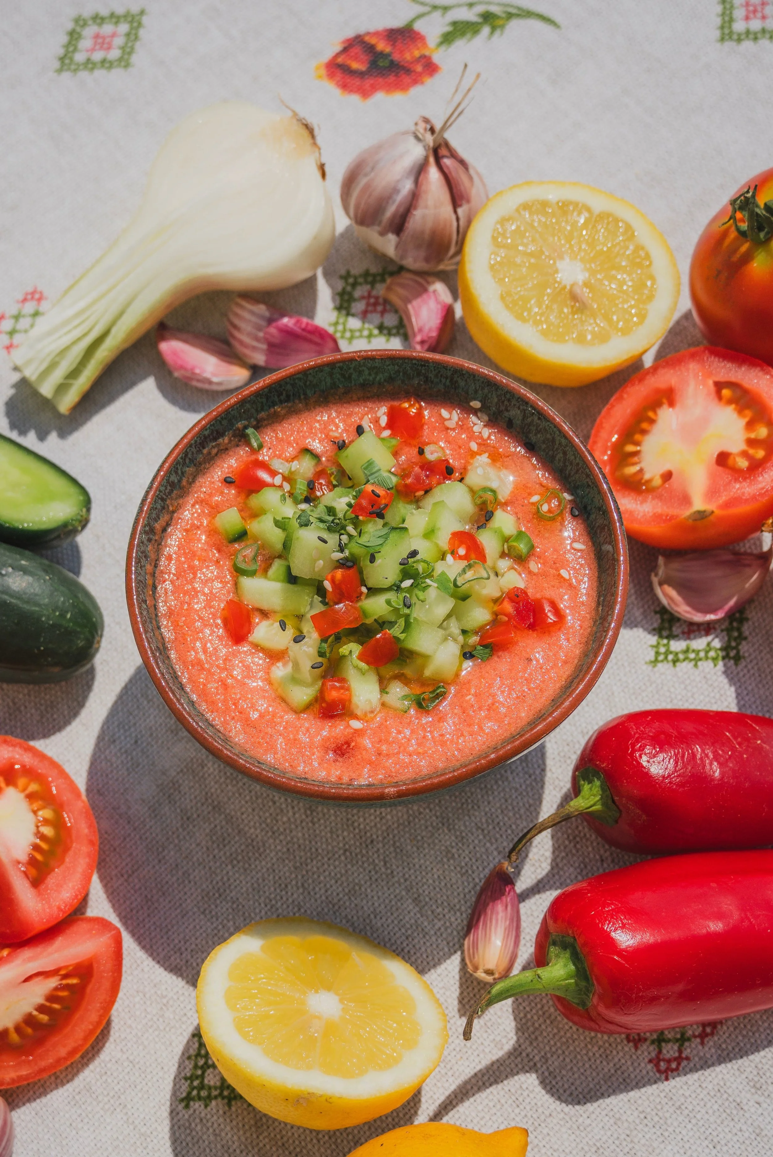 A bowl of cold soup garnished with chopped cucumbers and peppers, surrounded by fresh vegetables including garlic, lemon, tomatoes, cucumbers, red peppers, and jalapenos on a tablecloth.