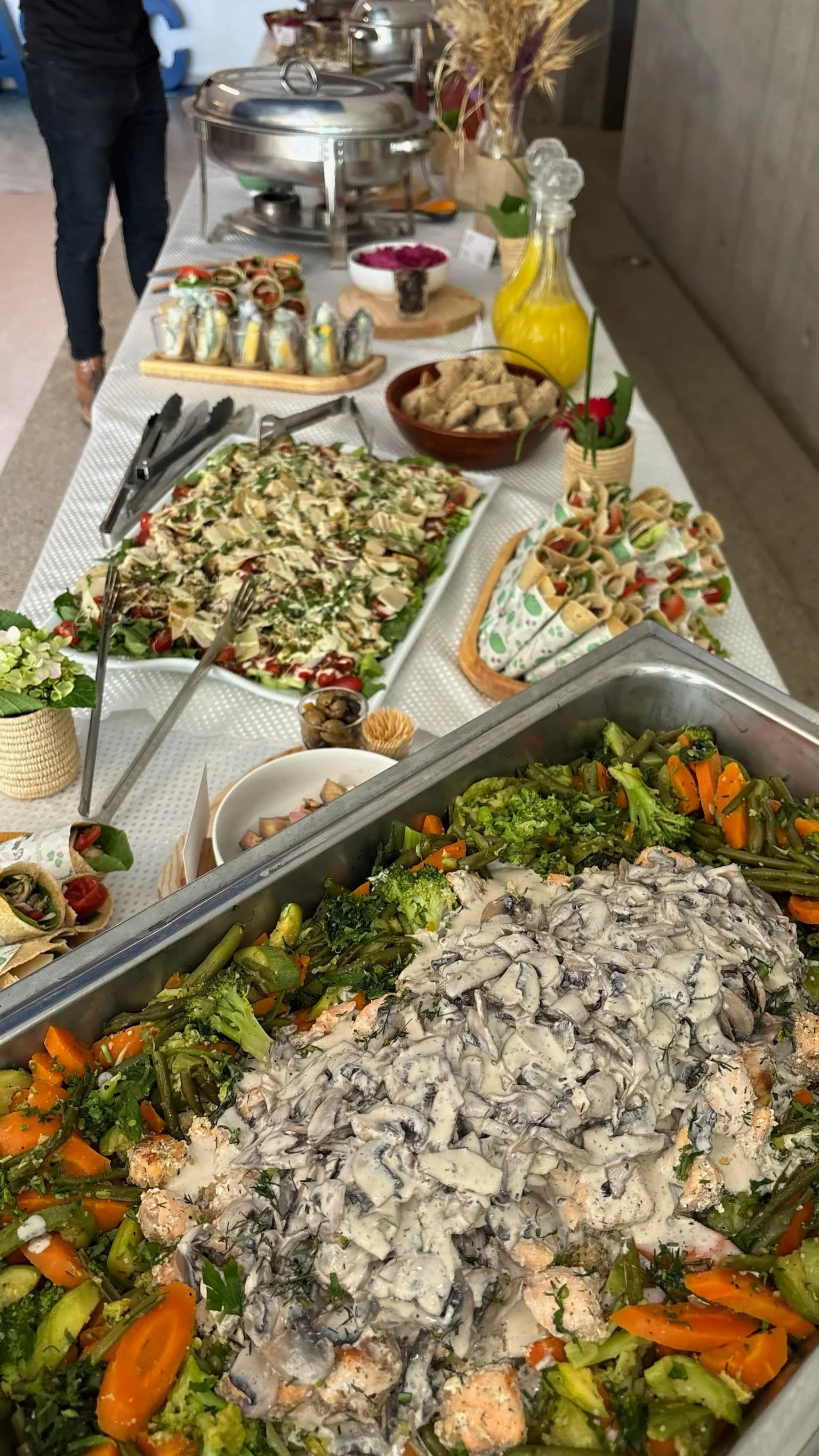 Buffet table with assortment of salads, vegetables, and finger foods including a large tray of mixed vegetables with a mushroom sauce, rolled wraps, a green and white salad, bread slices, and small appetizers, all arranged for a meal or event.