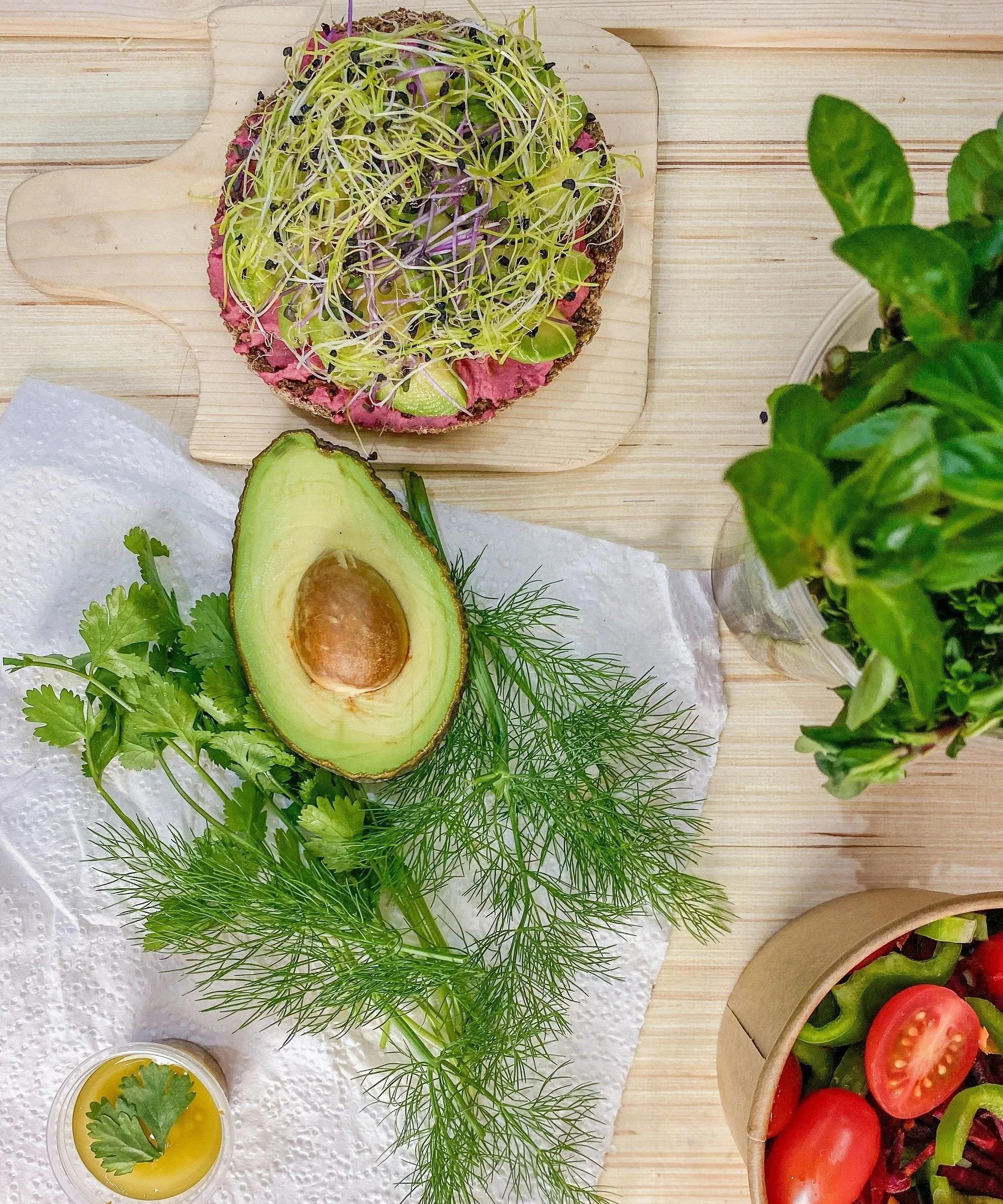 A sliced open avocado, fresh herbs, a small cup of oil with a cilantro leaf, a top view of a salad with cherry tomatoes, and an open-faced sandwich with sprouts and vegetables on a wooden surface.