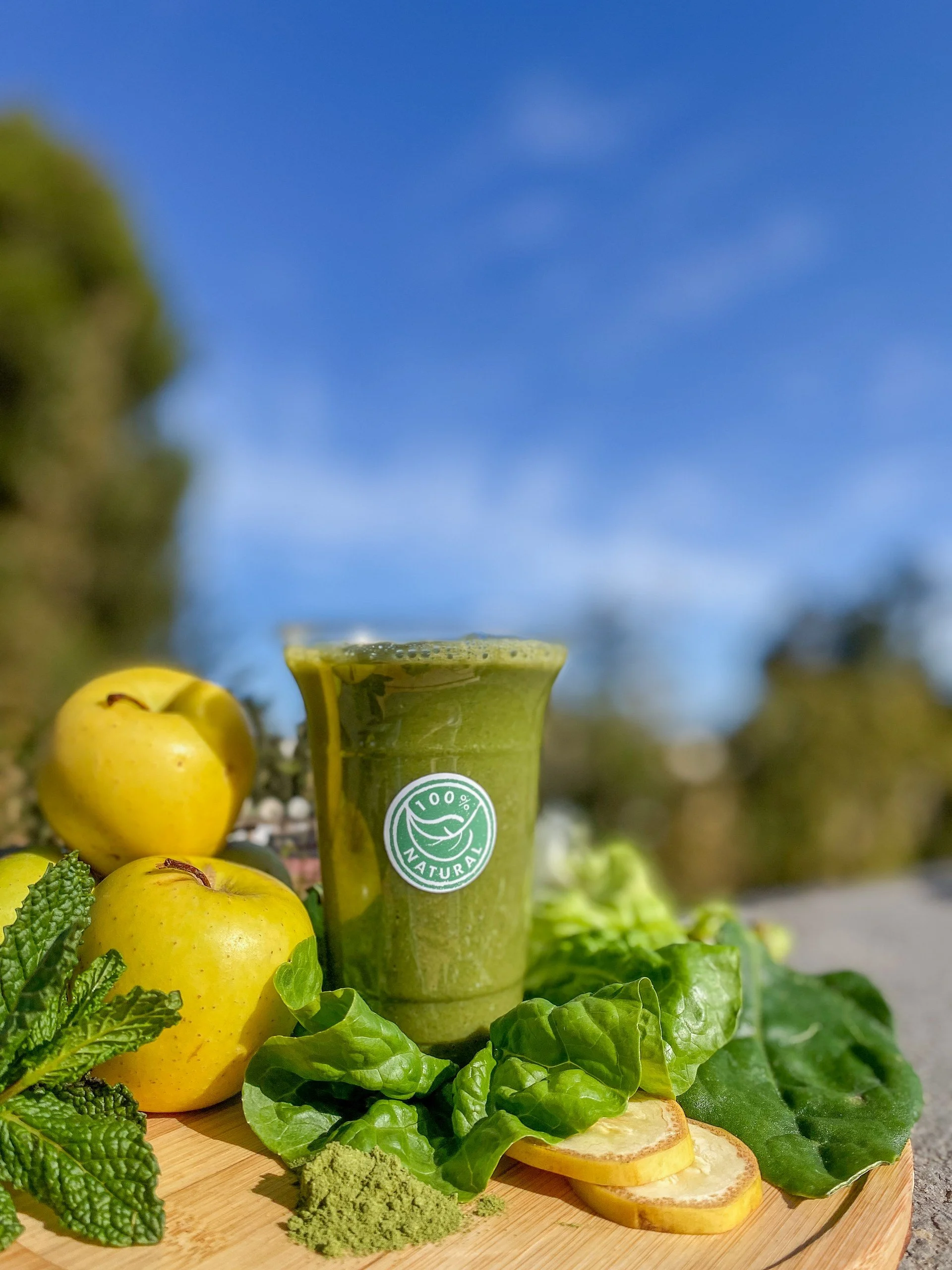A green smoothie in a clear glass with a '100% Natural' label, surrounded by yellow apples, fresh green lettuce and spinach, lemon slices, mint, matcha powder, and wheatgrass on a wooden surface outdoors with a blue sky in the background.