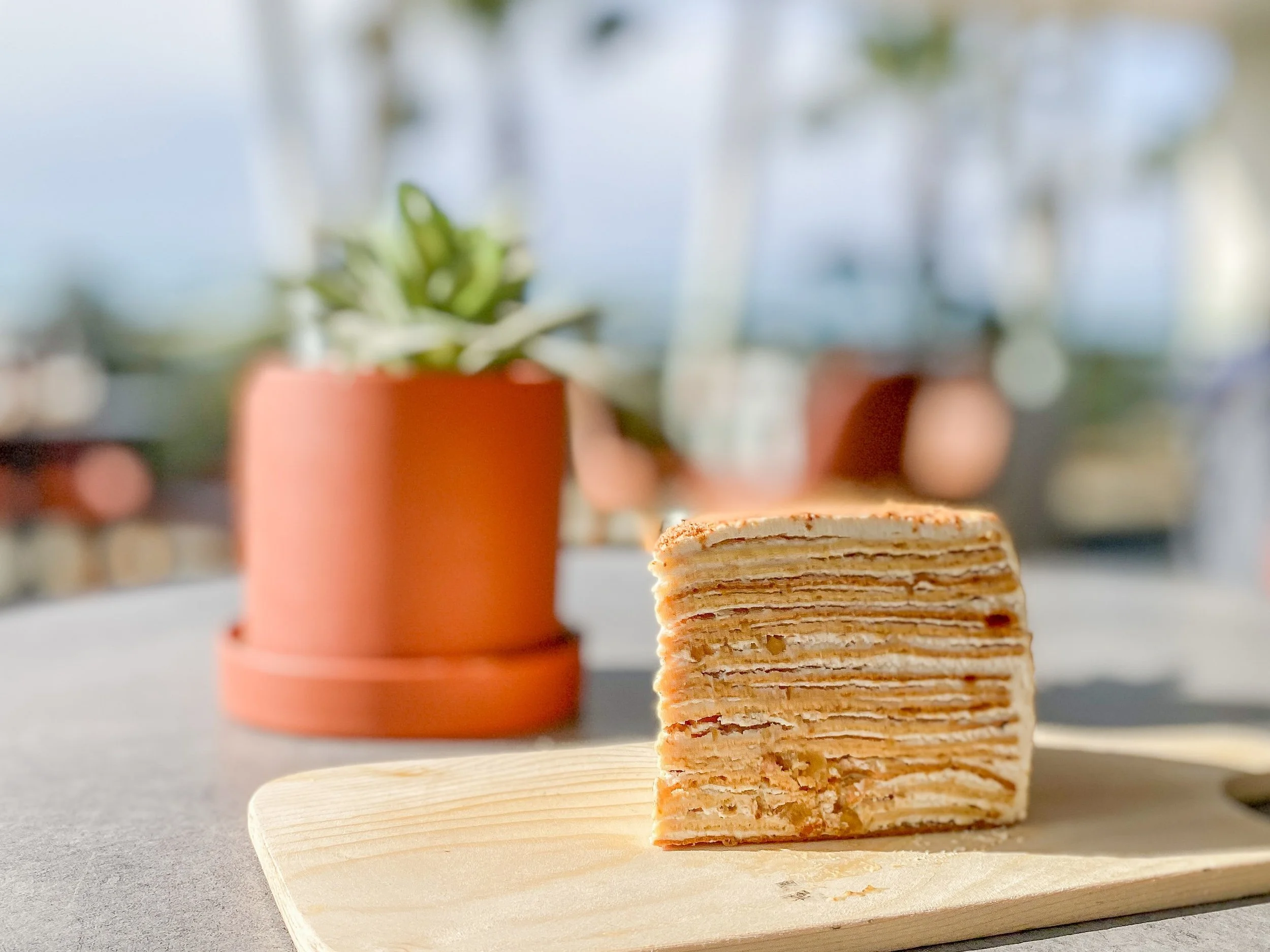 A layered slice of crepe cake with visible filling layers on a wooden board, with a blurred potted succulent and a bright window in the background.
