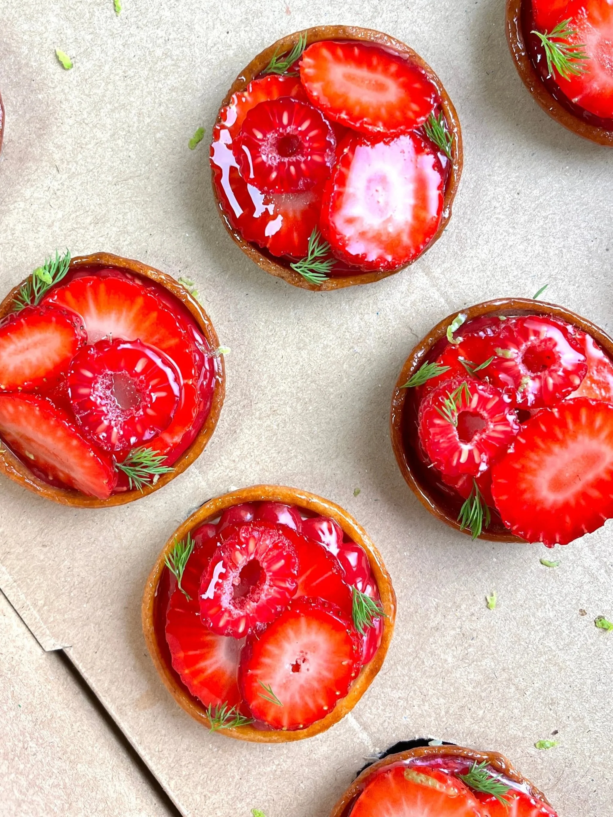 Strawberry tartlets topped with sliced strawberries and garnished with small sprigs of green herbs, arranged on parchment paper.