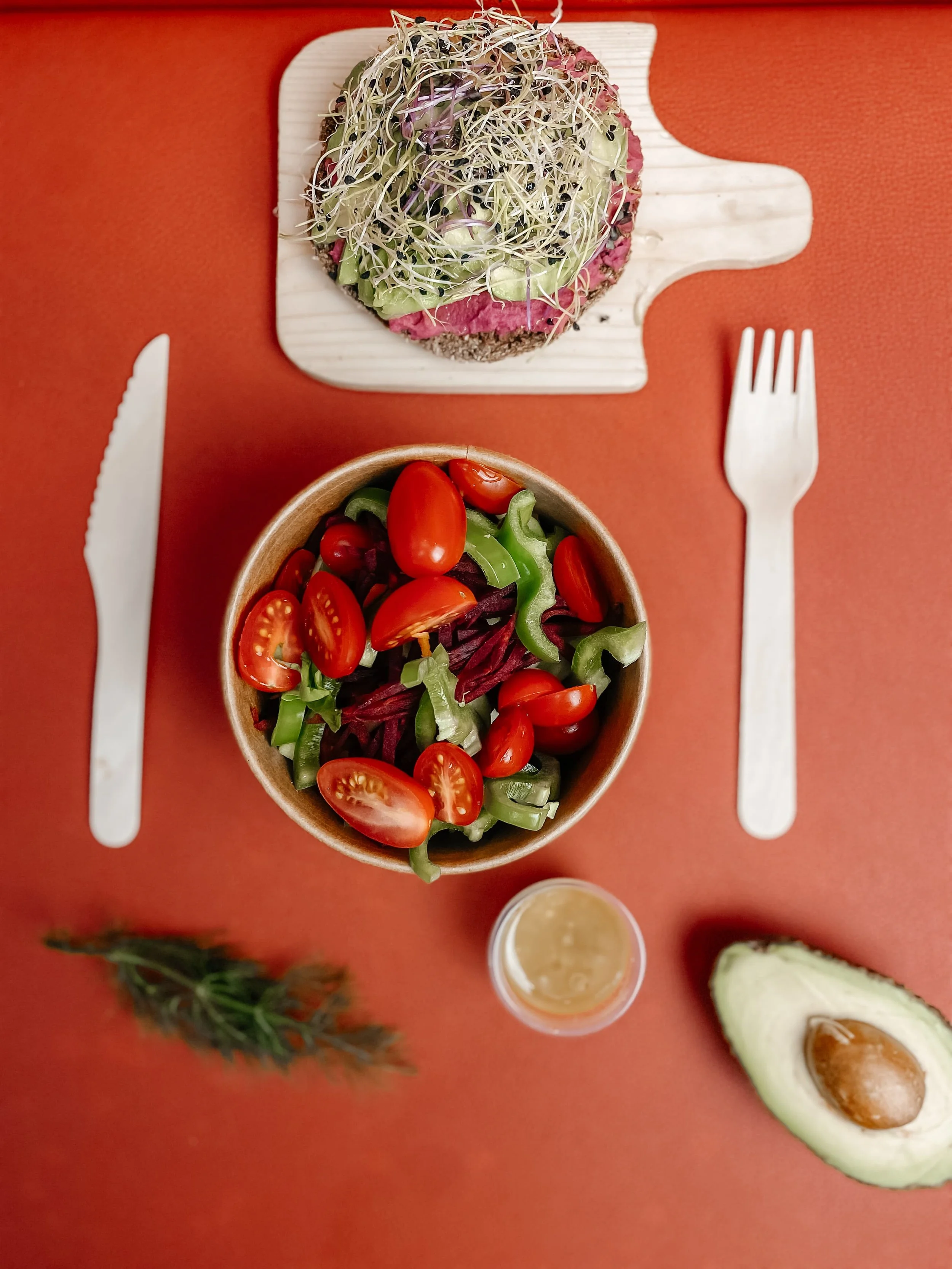 A wooden bowl filled with cherry tomatoes and sliced green bell peppers, a round plate with a round piece of toast topped with avocado, shredded sprouts, and olives, a halved avocado with the seed, a small cup with a yellowish drink, a sprig of fresh