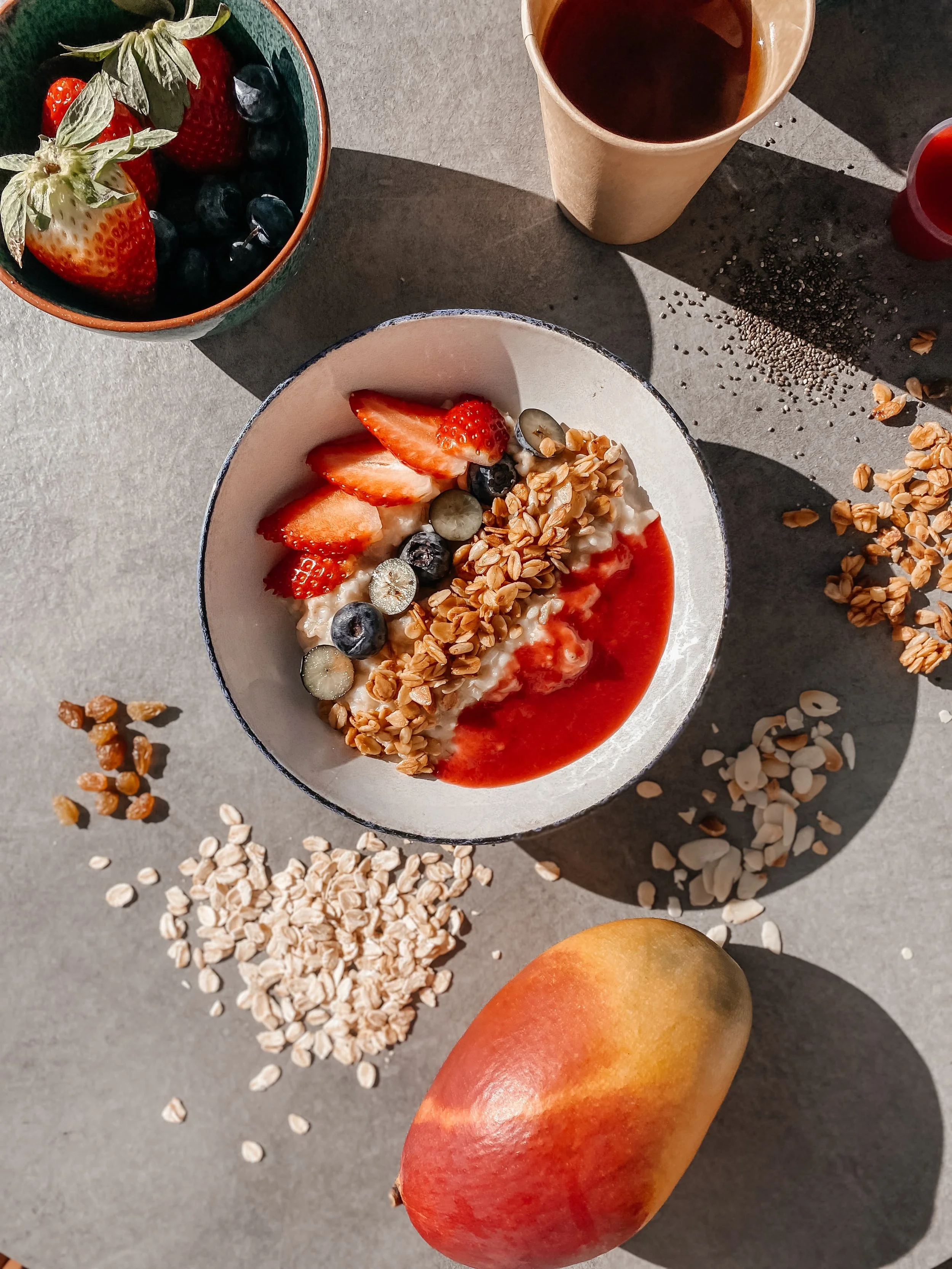 A top-down view of a breakfast scene with a bowl of yogurt topped with strawberries, blueberries, grapes, granola, and strawberry sauce, a cup of coffee, a small cup of berry juice, a bowl of fresh strawberries and blueberries, and a whole mango on a