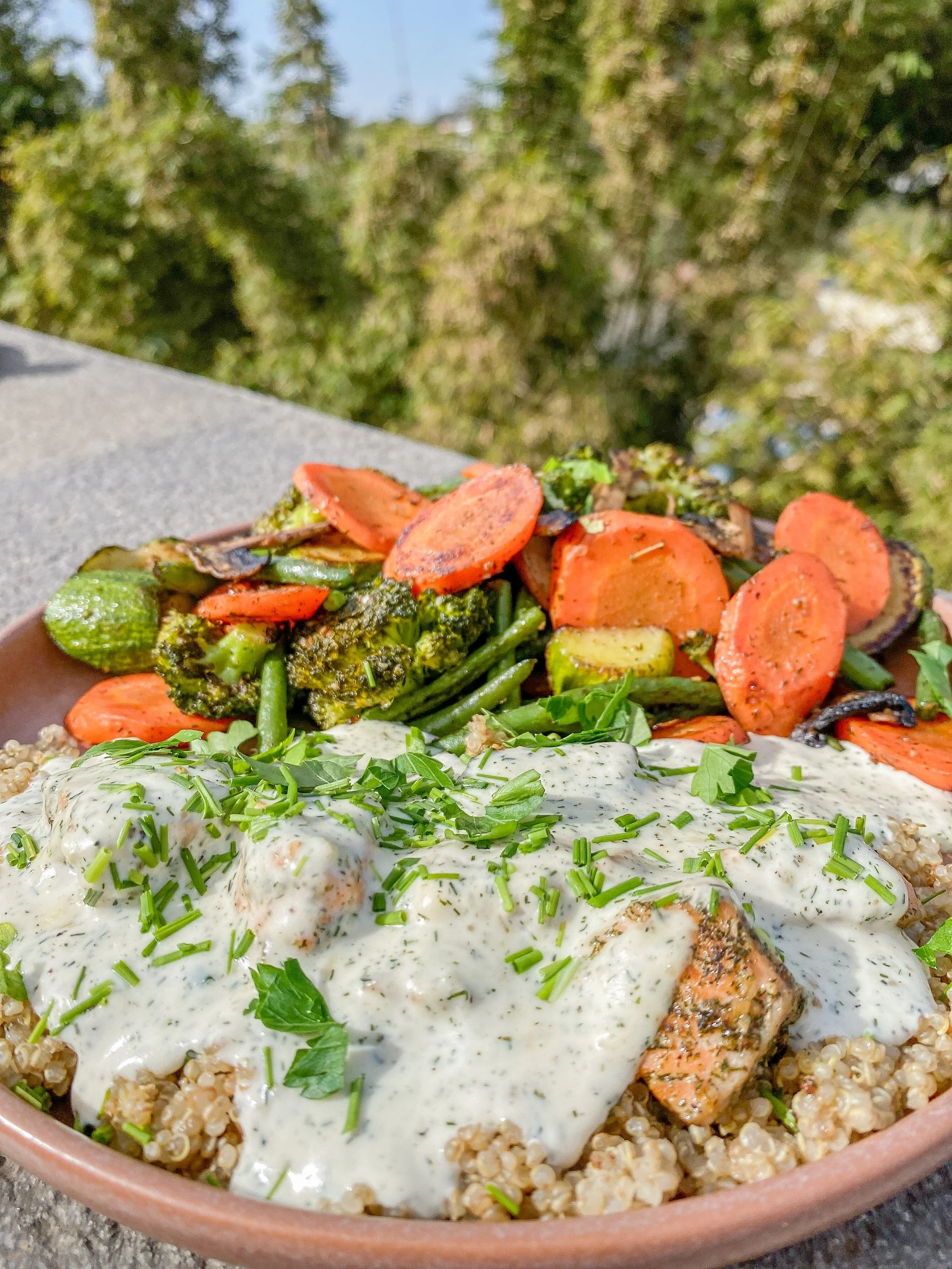 A bowl of cooked quinoa topped with cream-based herb sauce, grilled salmon, and garnished with chopped fresh parsley and chives, served with roasted mixed vegetables including carrots, broccoli, green beans, and eggplant.