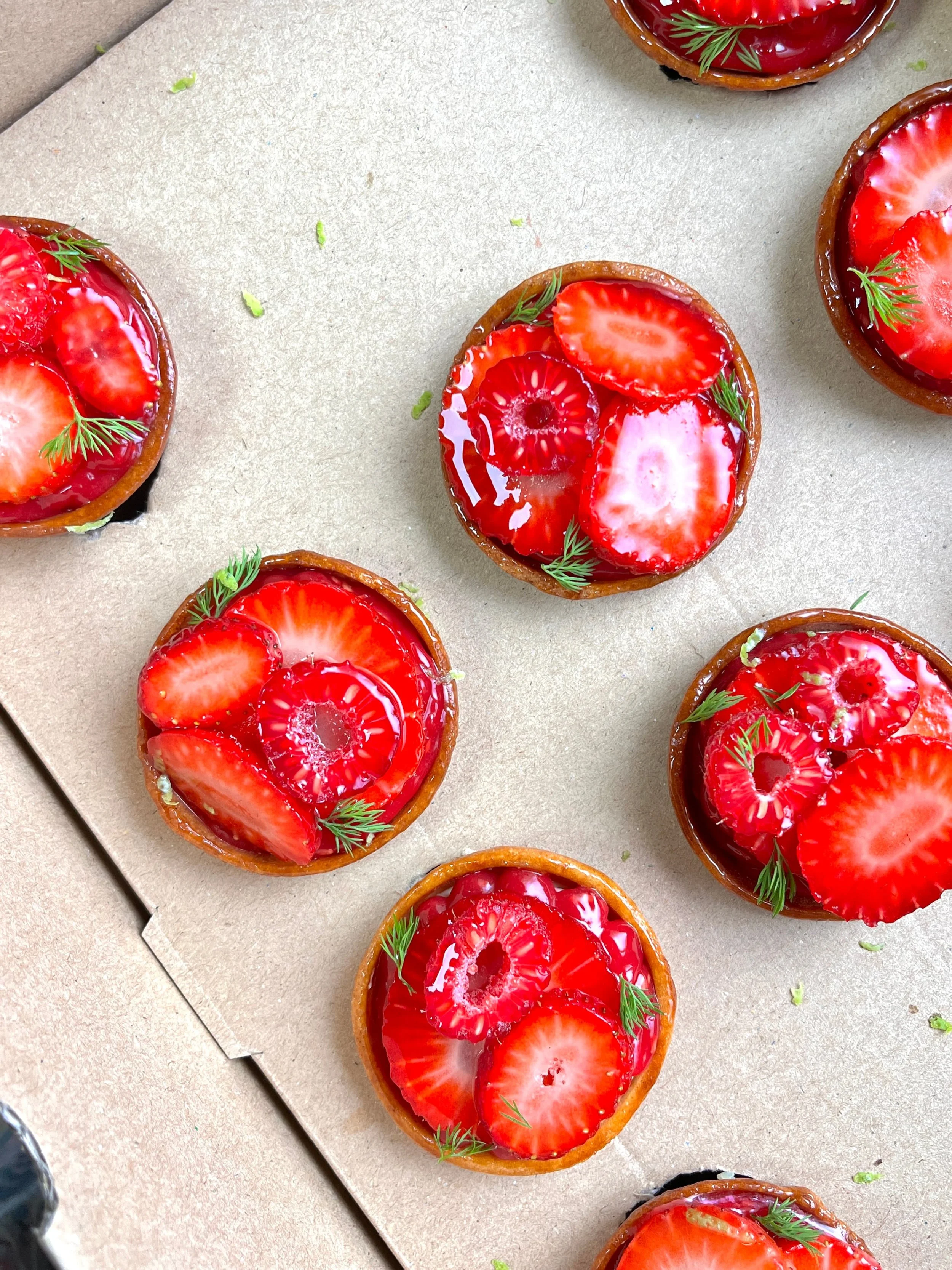 Small fruit tarts topped with sliced strawberries, red cherries, and small green herbs on a parchment paper.