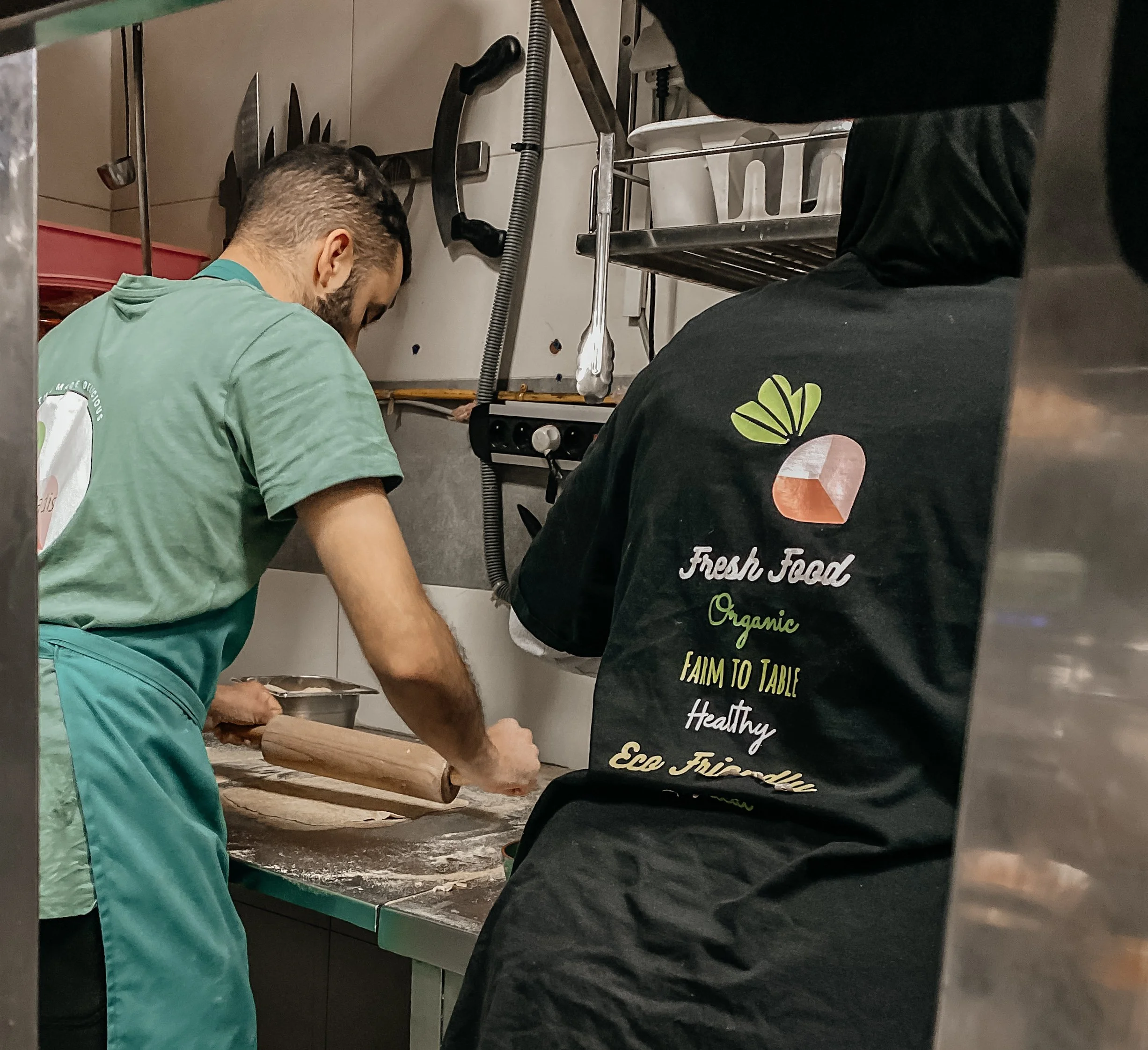Two people working in a kitchen, with one rolling dough on a green table and the other wearing a black T-shirt with food-related words and designs on the back.