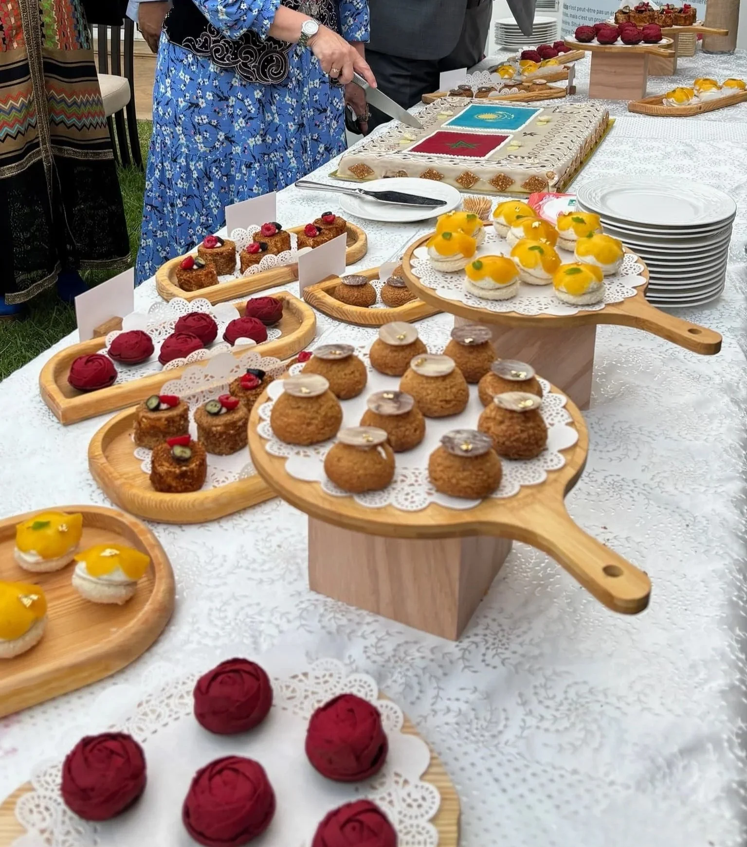 A variety of decorated pastries and desserts are displayed on wooden and paper platters on a table at an outdoor event, with a woman in a blue floral dress cutting a cake in the background.