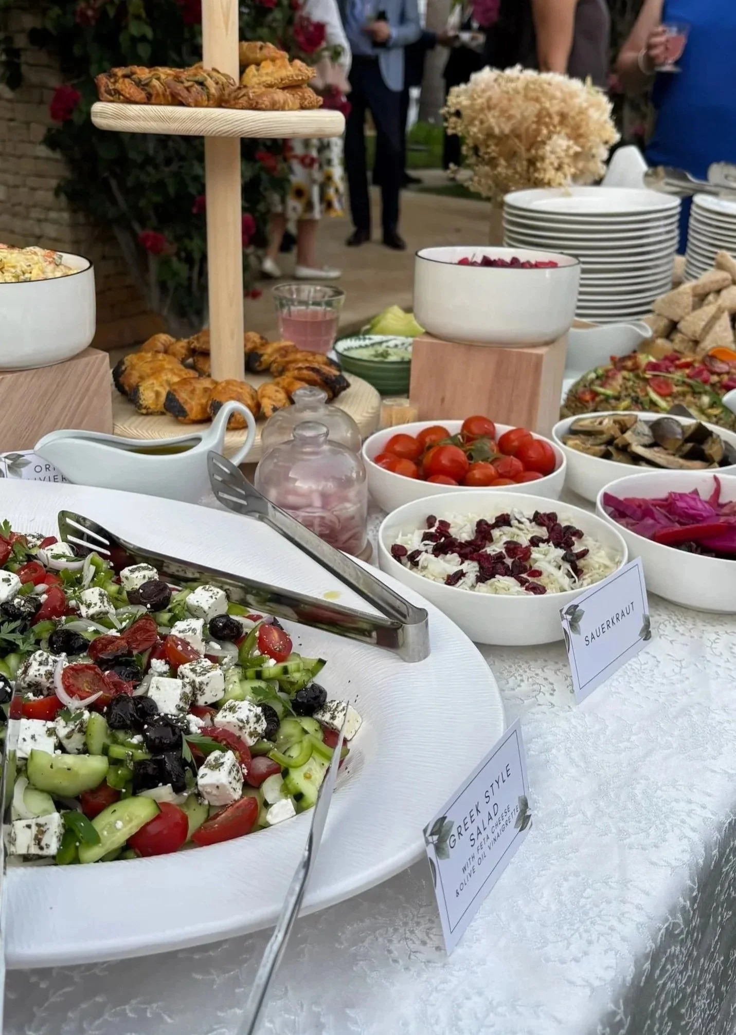 Variety of bowls and plates with salads, cherry tomatoes, pickled vegetables, and grilled chicken pieces on a buffet table at an outdoor event.