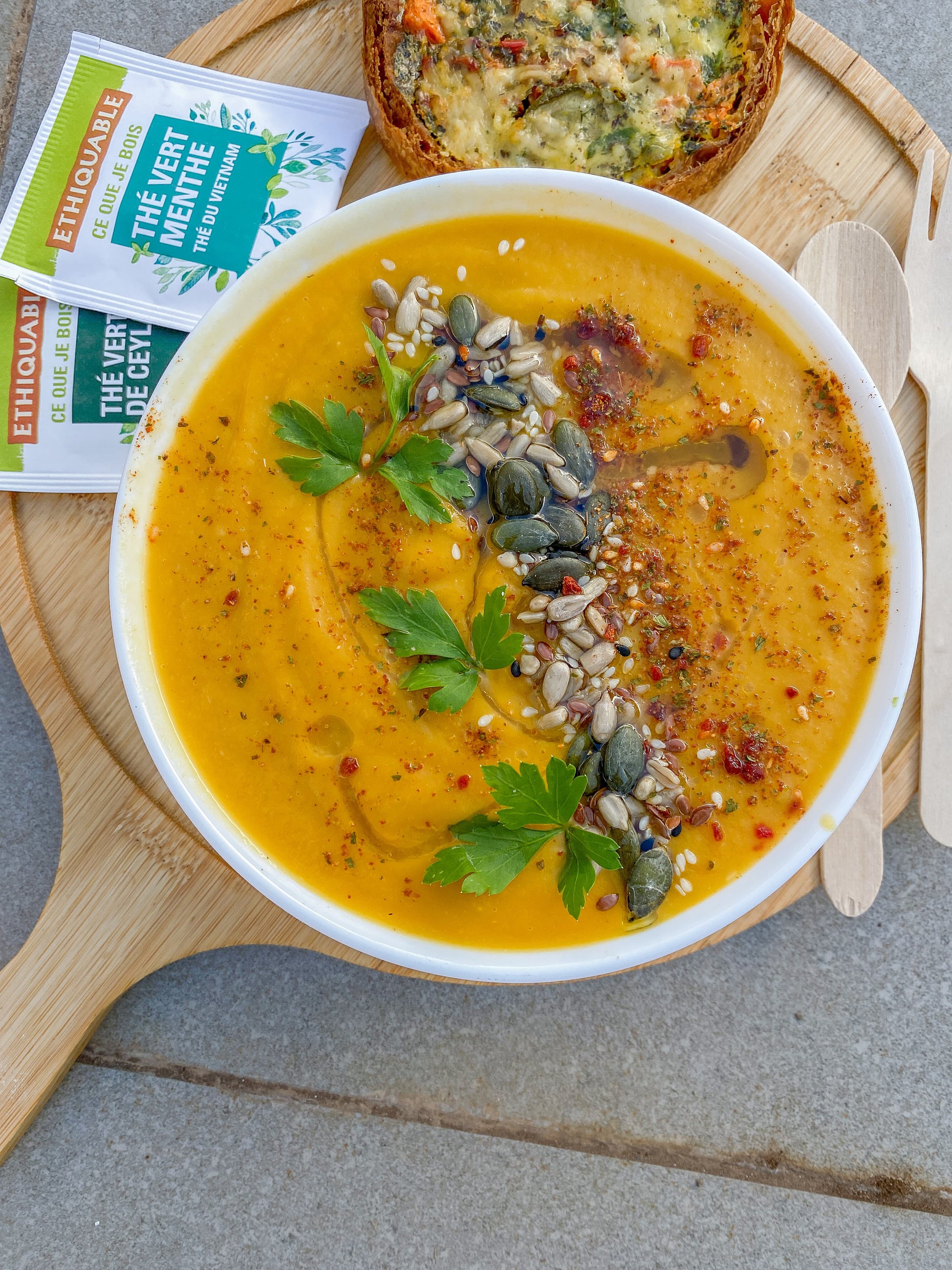 A bowl of bright yellow soup garnished with pumpkin seeds, sesame seeds, red pepper flakes, and fresh parsley, served on a wooden tray with a slice of vegetable quiche and two tea bags labeled 'The Veggie Menthe'.