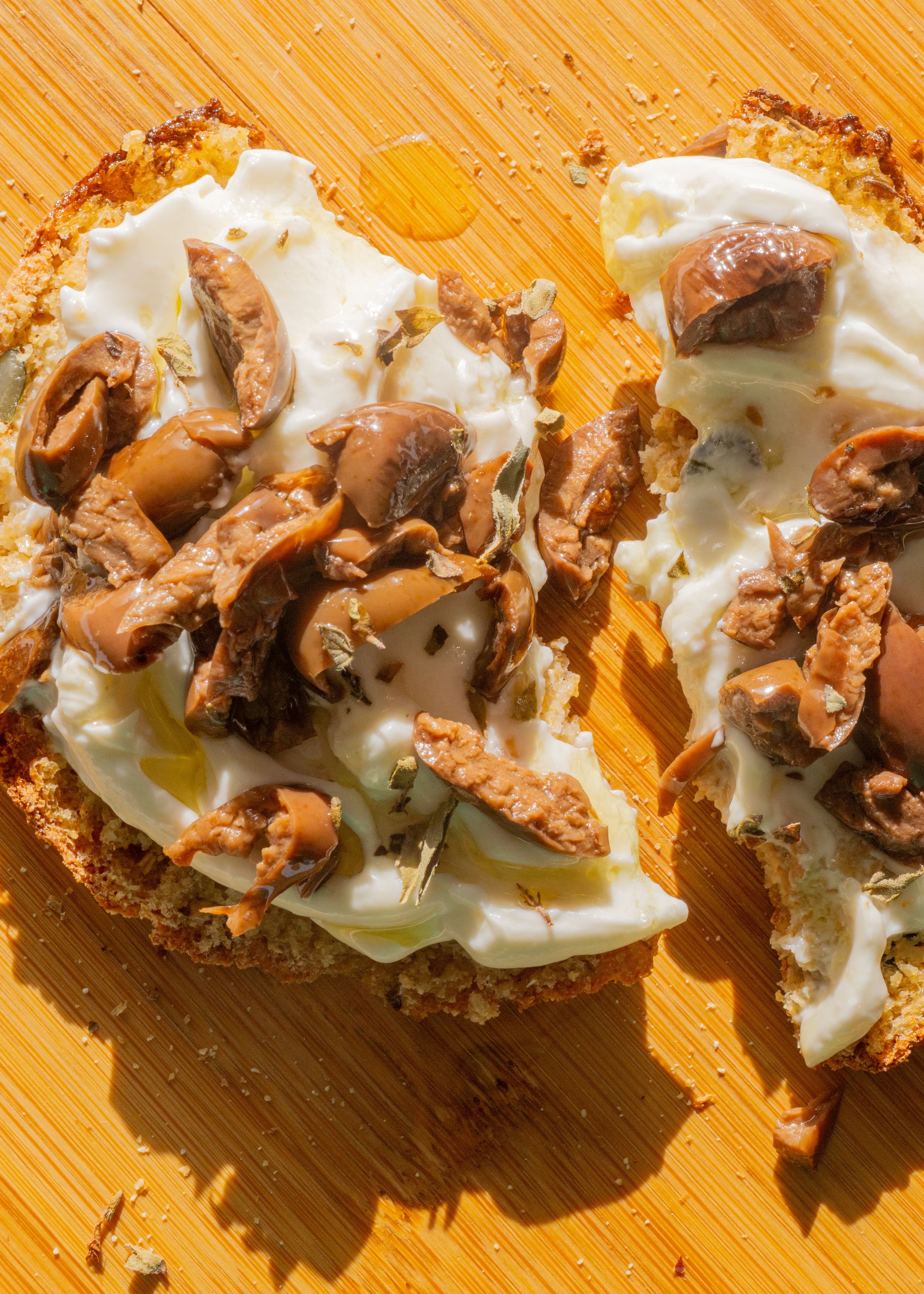 Slices of toasted bread topped with creamy yogurt, chopped olives, and herbs on a wooden cutting board.