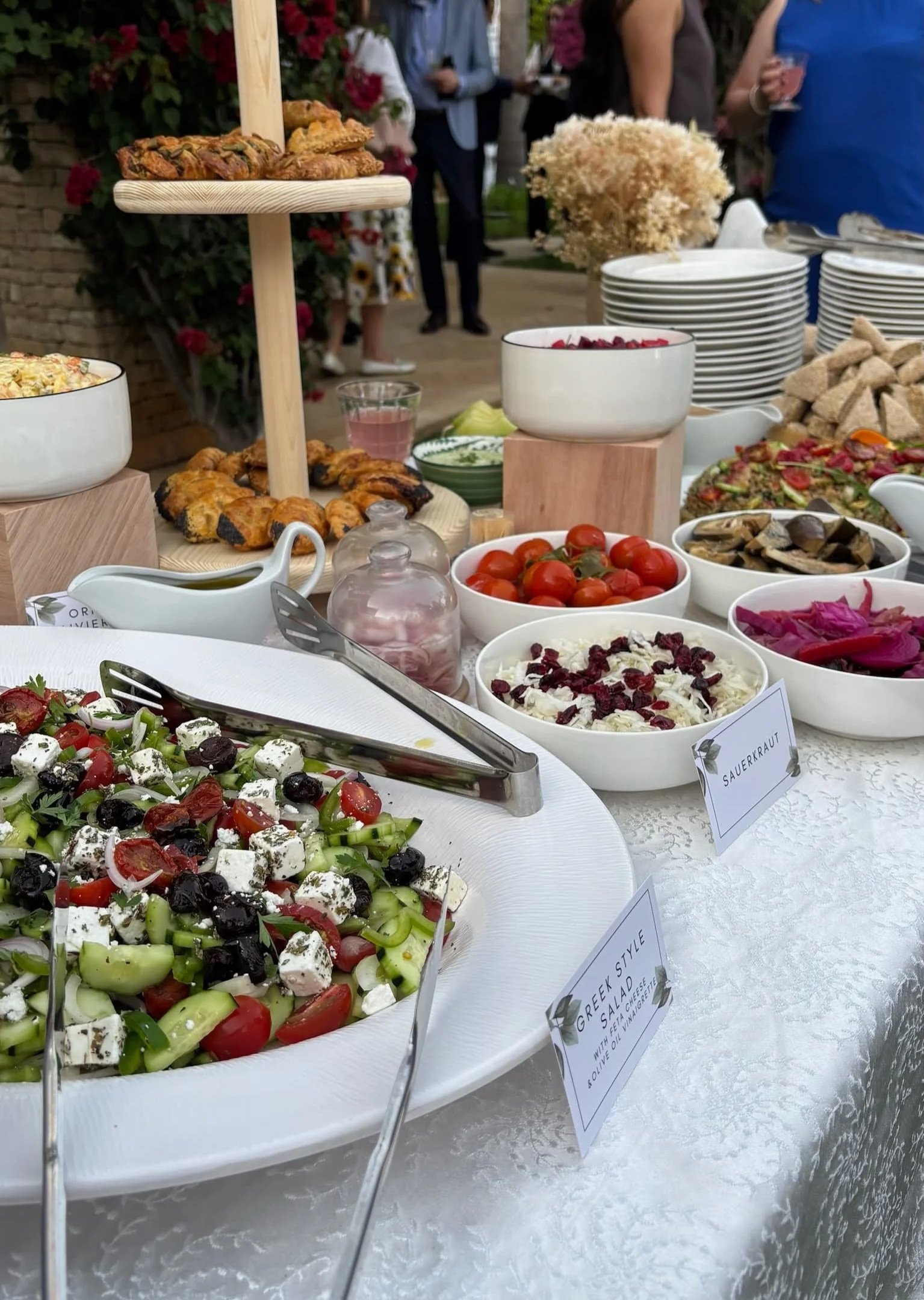 A buffet table with various salads, tomatoes, and baked goods at an outdoor event, with people socializing in the background.