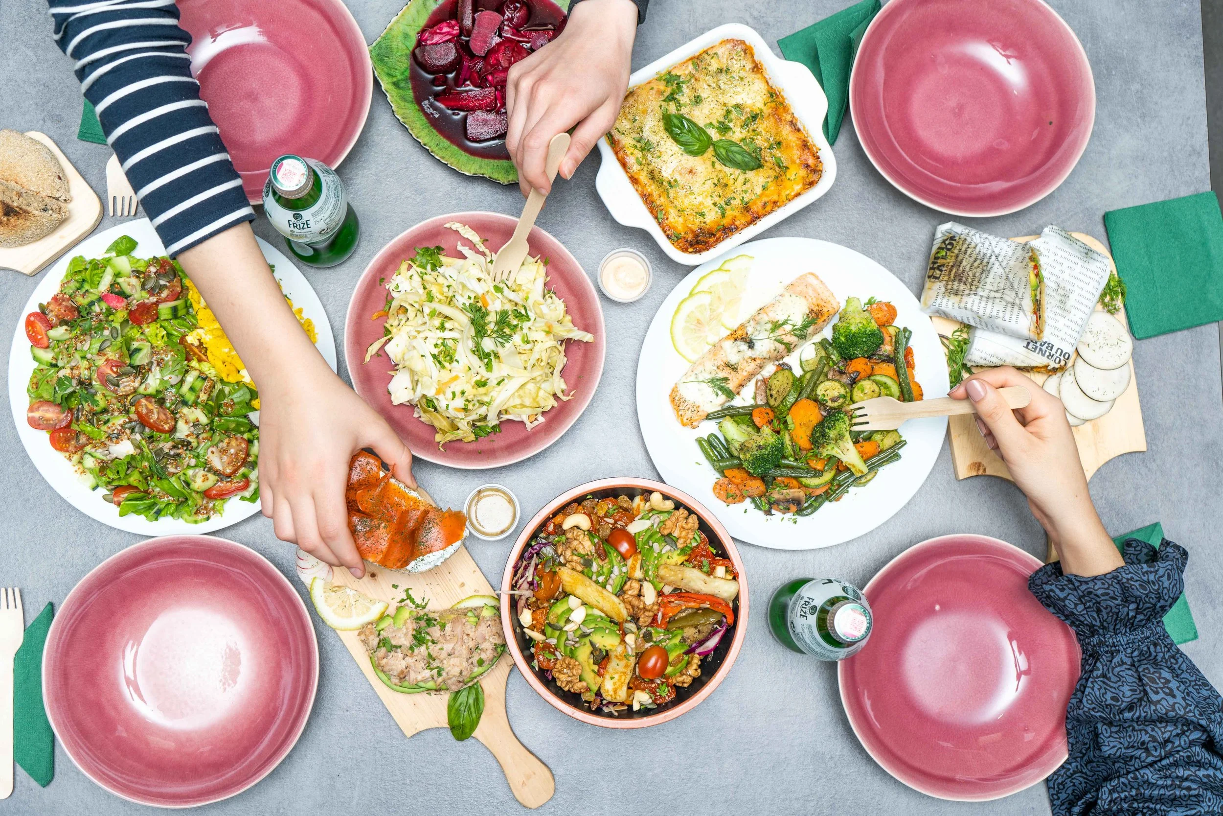 A table filled with various dishes including salad, pasta, grilled salmon, roasted vegetables, and beet salad, with hands reaching for food and pink plates arranged around.