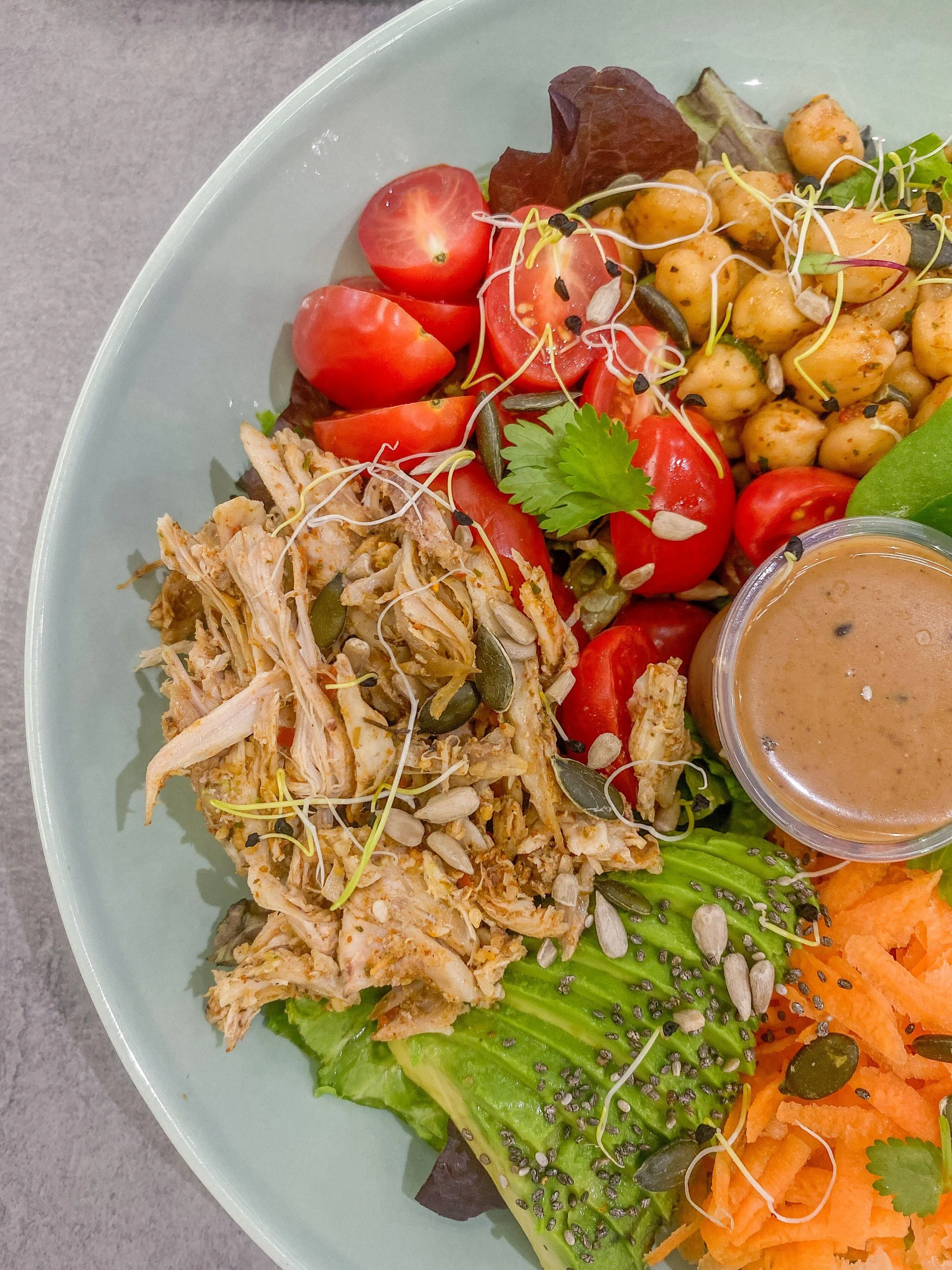 A colorful salad with cherry tomatoes, shredded chicken, chickpeas, shredded carrots, lettuce, avocado, sunflower seeds, sprouts, a cilantro leaf, and a small cup of salad dressing on a light green plate.