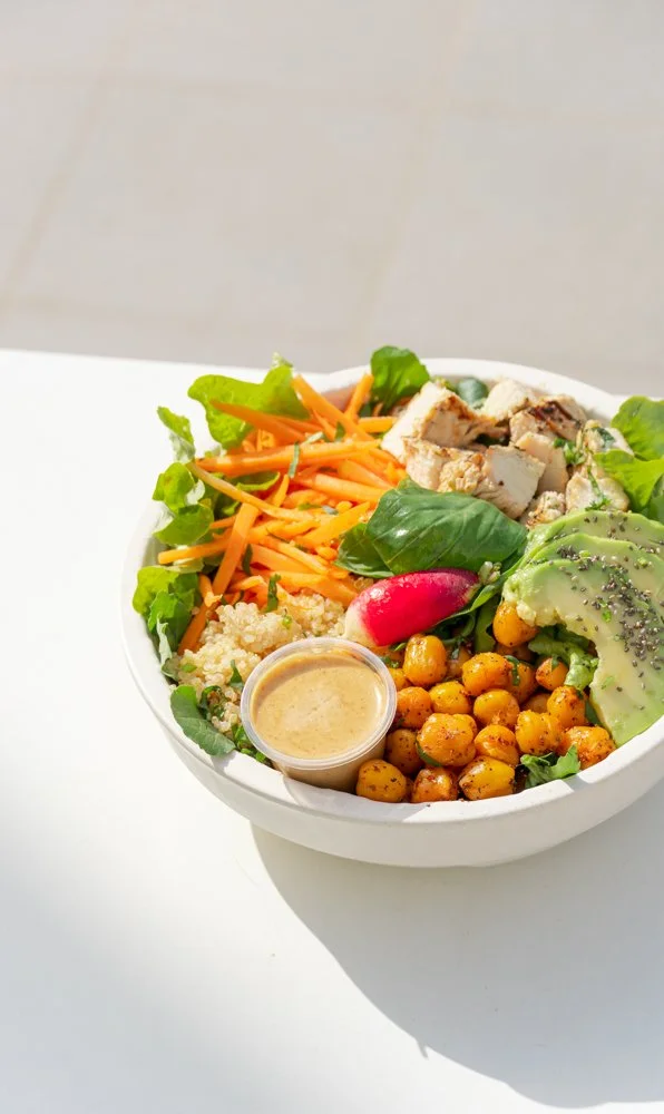 Salad bowl with shredded carrots, sliced chicken, leafy greens, radish, roasted chickpeas, avocado slices, and a small container of dressing.