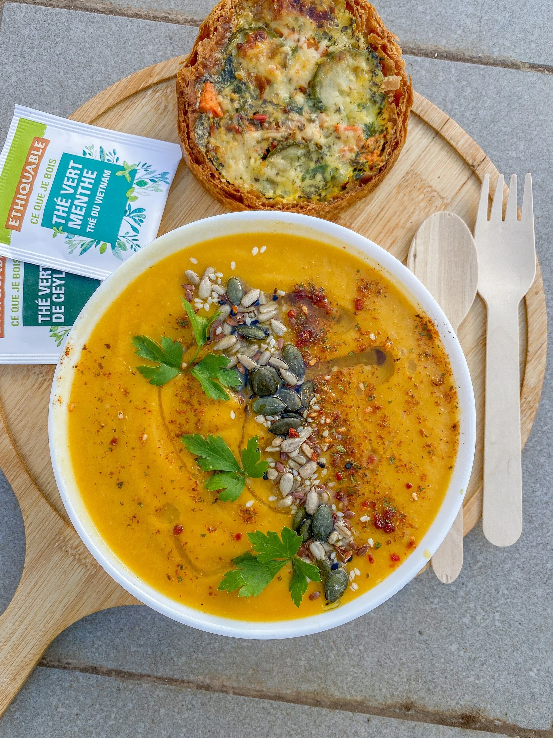 A bowl of yellow soup garnished with seeds and herbs, a slice of bread with vegetables and cheese, on a wooden tray with a wooden spoon and fork, and packets of tea.