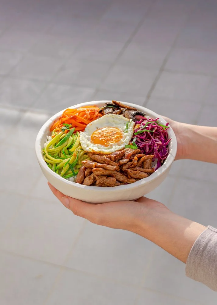 A bowl of assorted colorful ingredients including shredded carrots, zucchini noodles, sliced mushrooms, shredded purple cabbage, cooked meat, and a fried egg, being held by two hands.