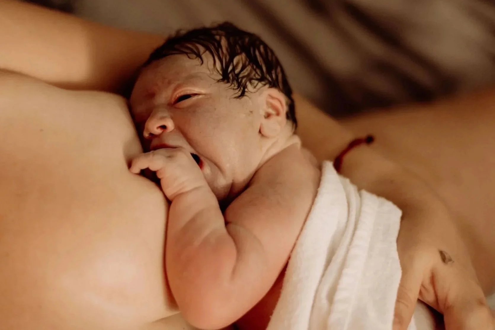 A white newborn baby lying skin to skin on their white parent's chest shortly after birth. The baby is partially covered with a white blanket.