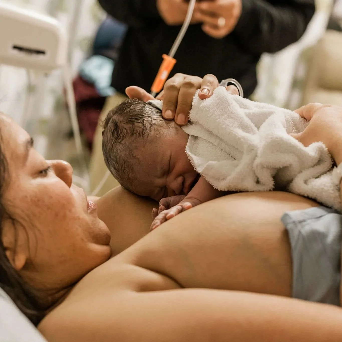 A Black femme presenting person tenderly holding their baby skin to skin immediately after their C-section birth. Their partner stands in the background, using their phone.