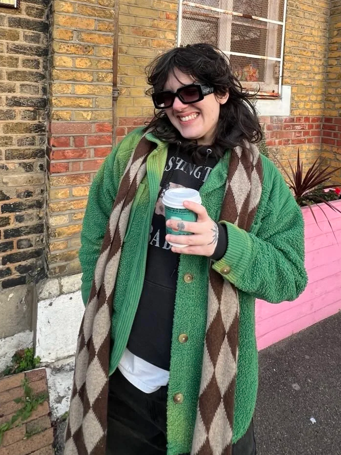 Joey is a white non-binary person, they are smiling and holding a coffee cup outside, wearing black sunglasses, a green cardigan, a black hoodie, and a brown and beige diamond-patterned scarf, with a brick wall and pink planter in the background.