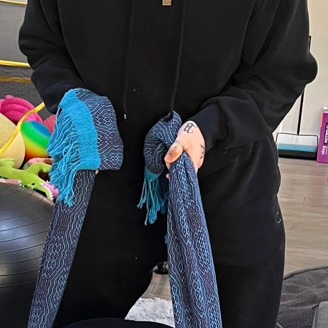 A close up shot of Joey's torso, arms and legs while they perform sifting with a blue and purple rebozo, their pregnant client is just out of sight. Joey is wearing a black tracksuit and gold thumb ring.