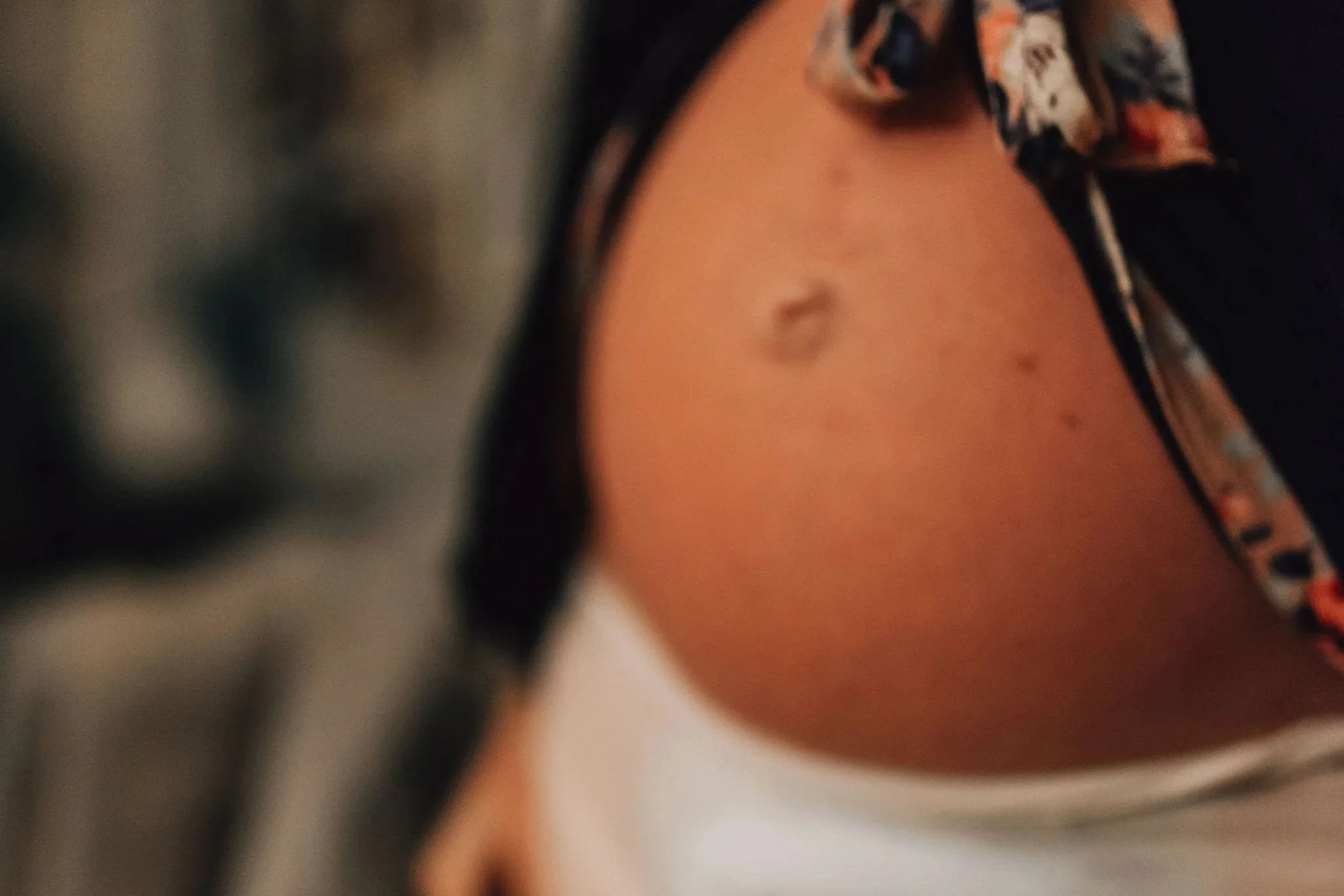 Close-up of a white pregnant person's midsection during labour at home, revealing their belly and one of their hands, they are wearing a white adult diaper and a black dressing gown with pink and red floral trim.