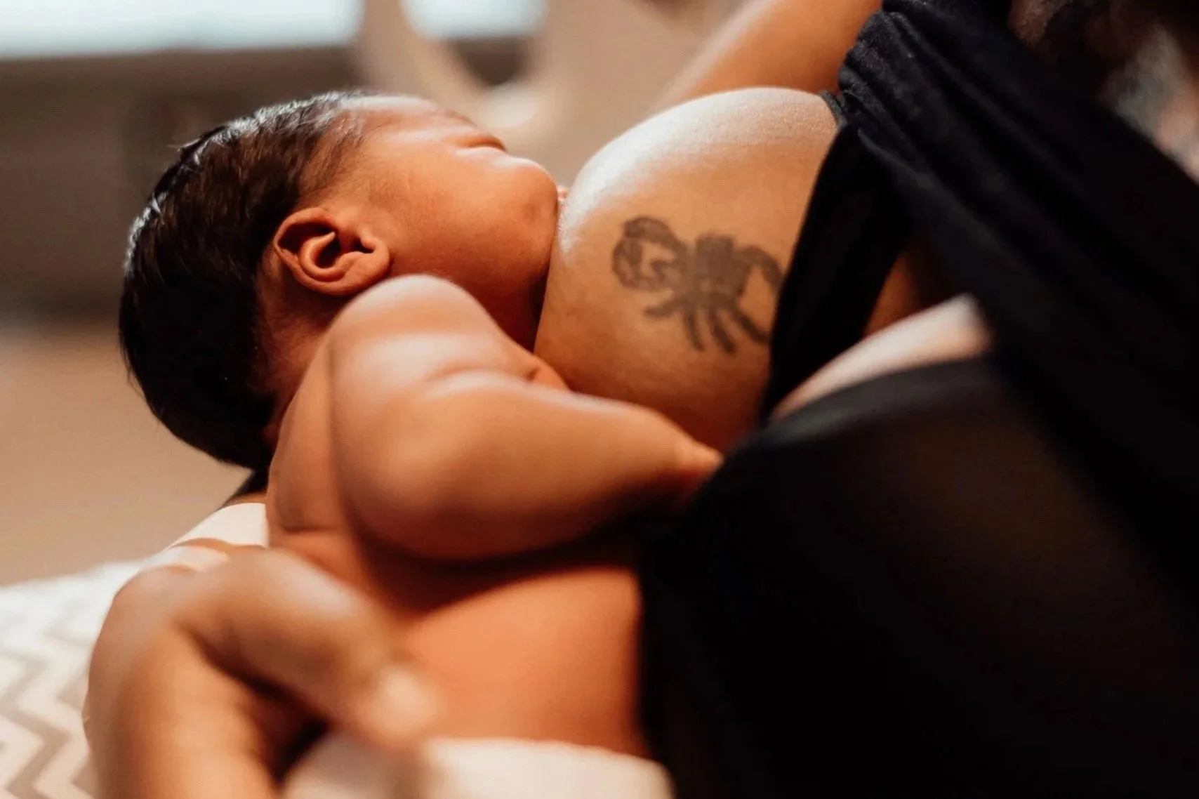 A Black newborn baby body-feeding from their Black parent who is wearing a black top, they have a tattoo on the side of their chest the baby is feeding from.
