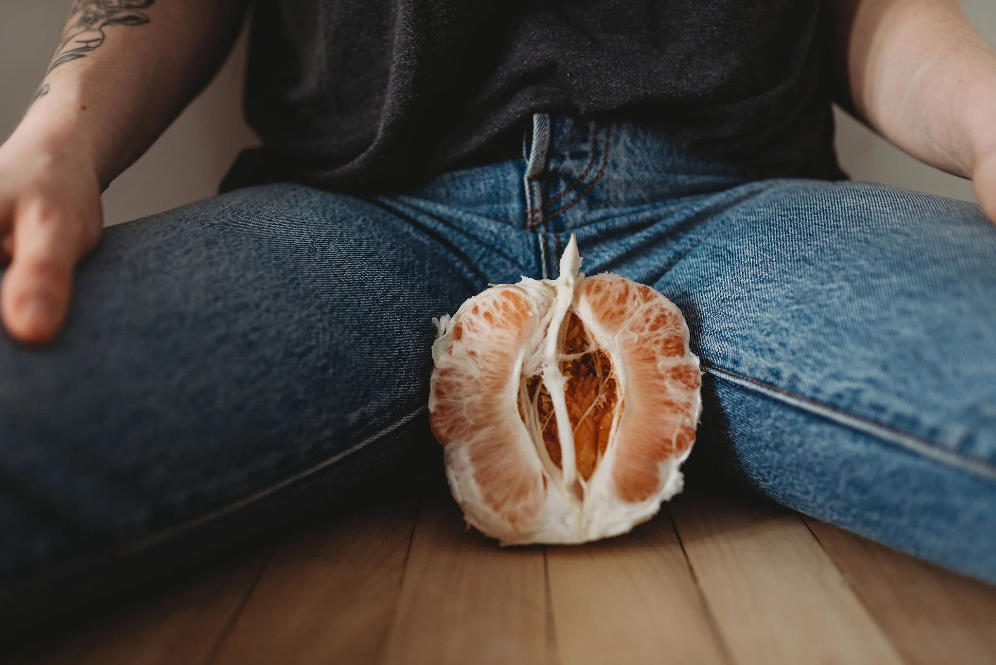 A person sitting on the floor with their legs spread, wearing blue jeans and a dark shirt, with a half a peeled grapefruit placed between their legs. The middle of the grapefruit looks similar to female genitalia.