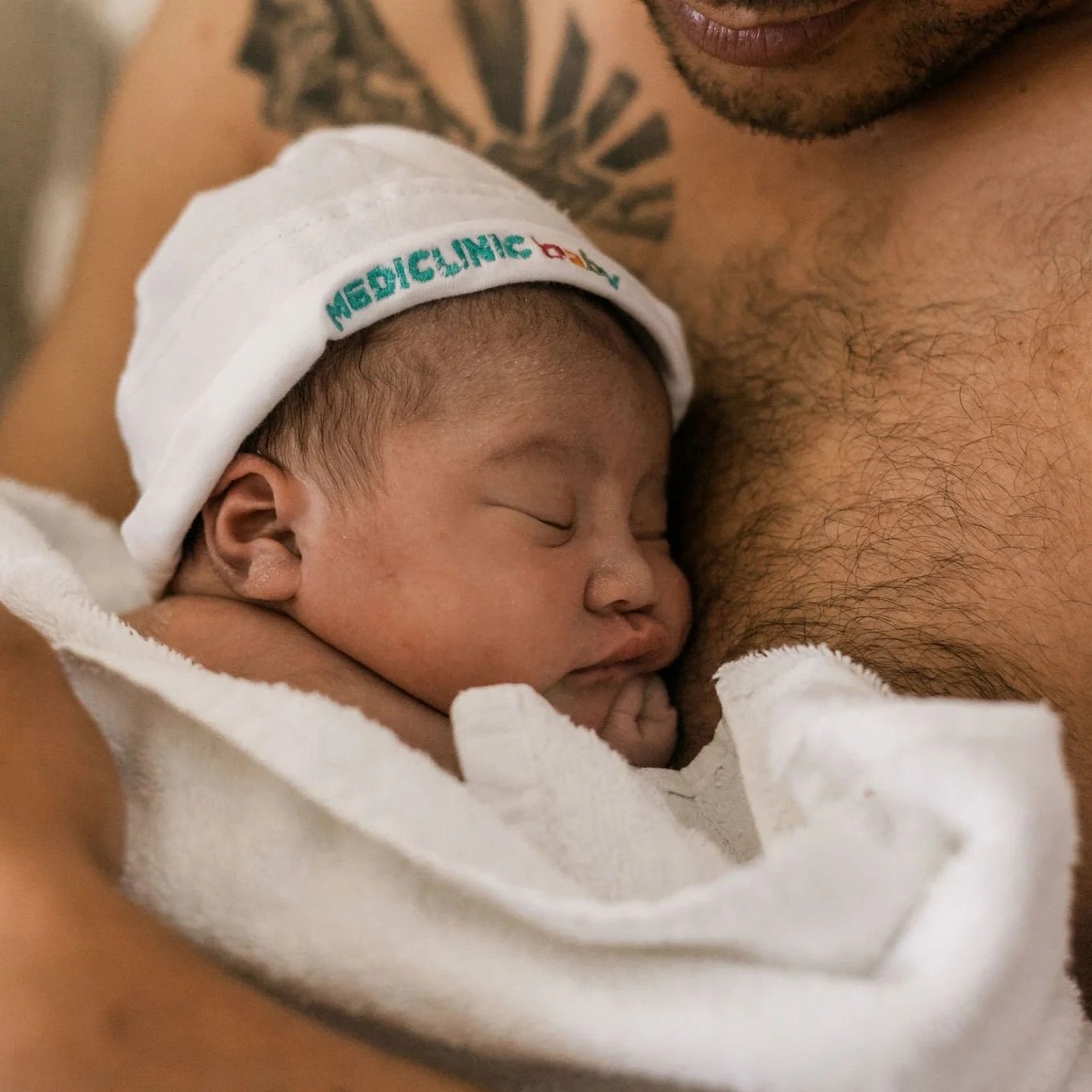 Close-up of a Brown newborn baby sleeping skin to skin on their Brown parent's bare chest, the baby is wearing a white hat with 'MEDICINE' written on it, and they are wrapped in a white blanket.