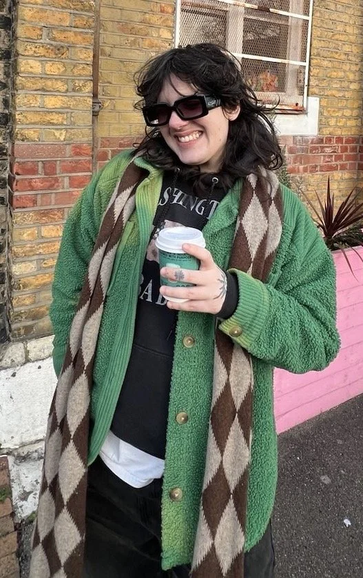 Joey is a white non binary person with dark brown hair. They are wearing a green cardigan, white tshirt under a black hoodie, jeans and a brown patterned scarf. They are wearing sunglasses and holding a green coffee cup in front of a brick wall
