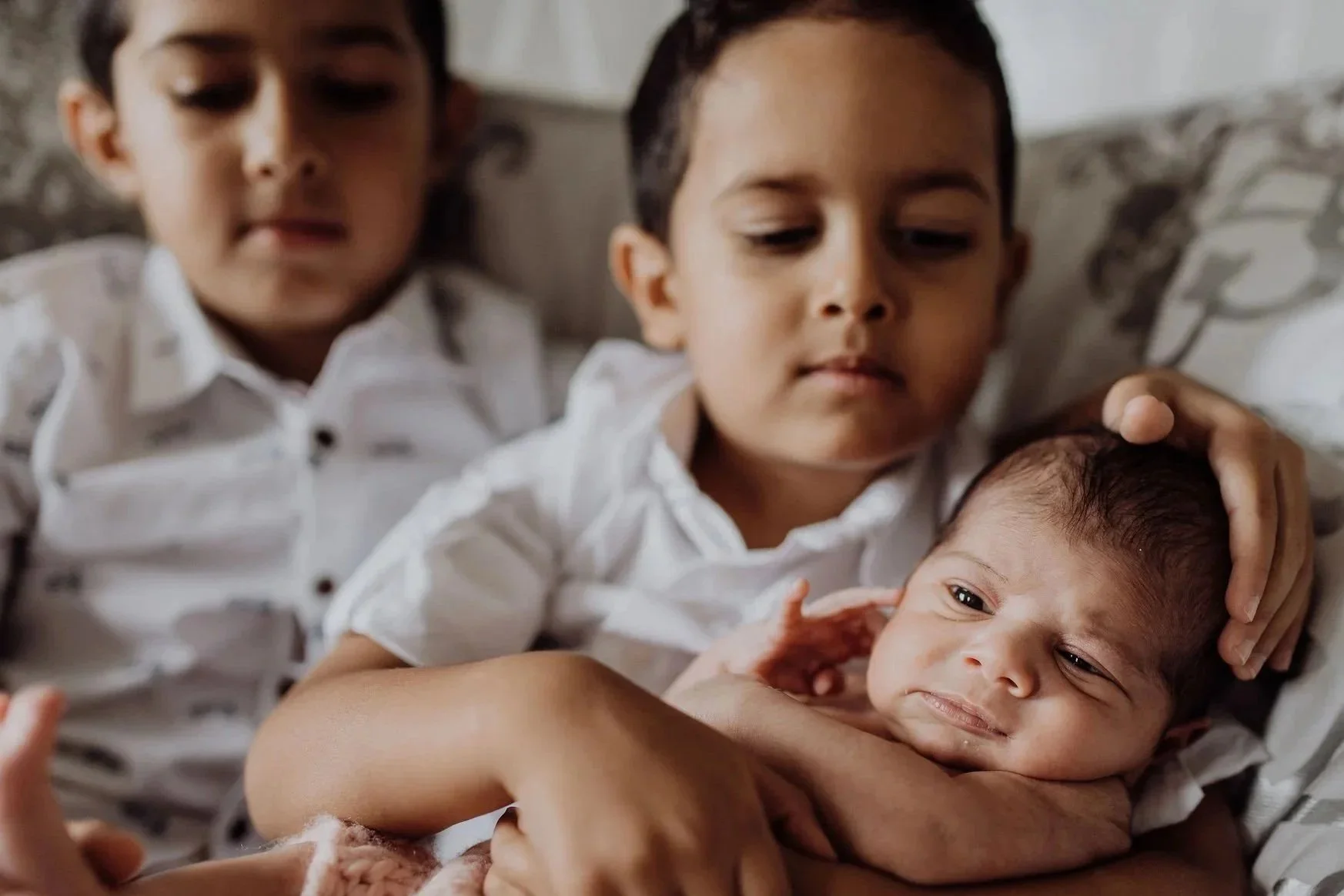 Two Brown siblings of child age, holding their Brown baby sibling, one of the children is stroking the baby's hair. The children are wearing white shirts and the baby is wearing a pink top.