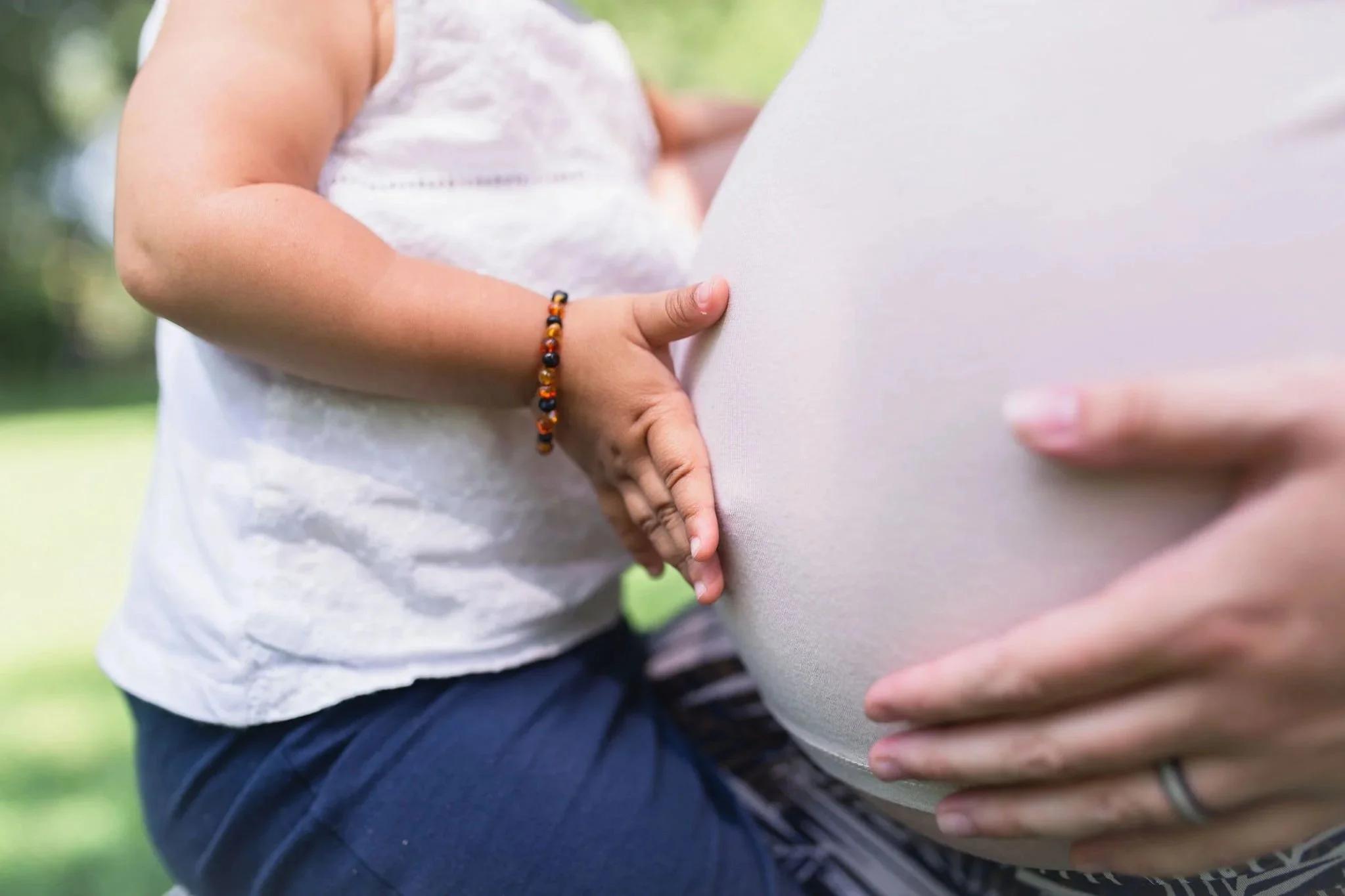 A white toddler touching their white parent's pregnant belly. They are both wearing white tops, the child has on a brown bracelet and blue jeans. The adult's hand is touching their belly.