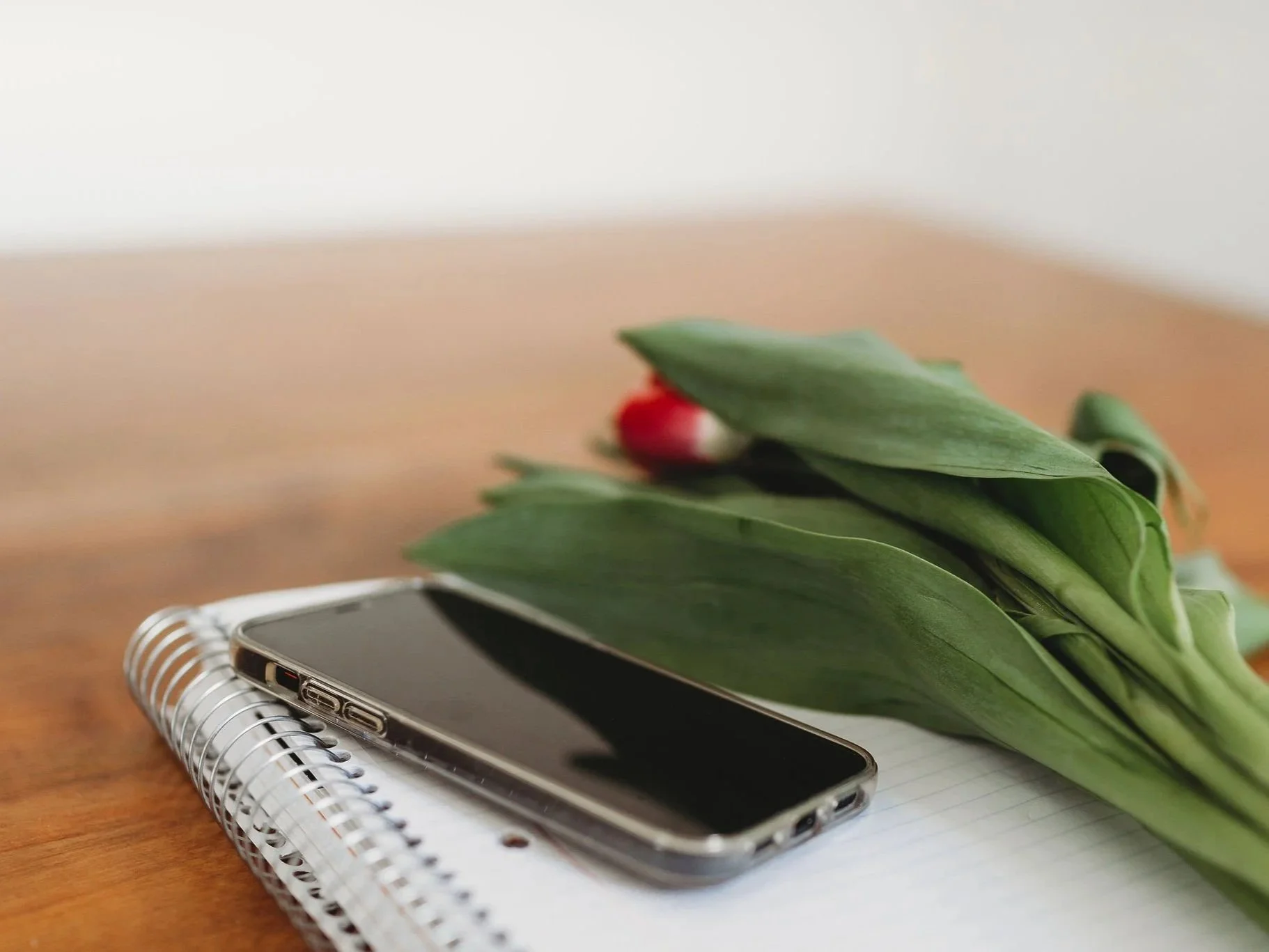 A smartphone, a spiral notebook, and a bouquet of tulips with green leaves on a wooden surface.