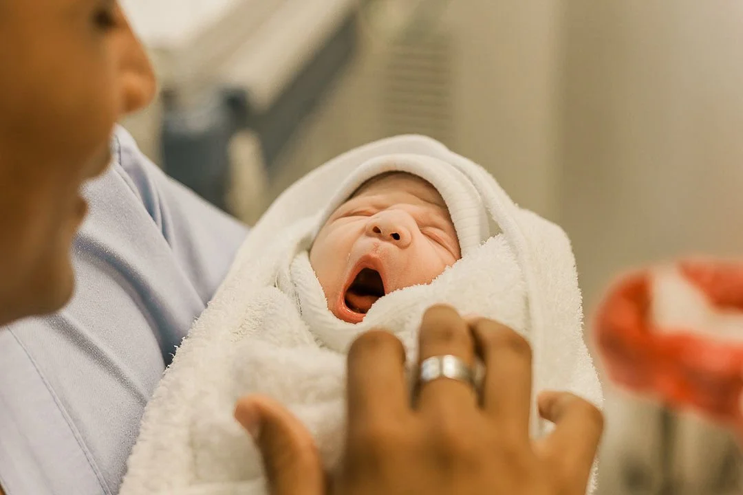 Close-up of a newborn baby wrapped in a blanket and wearing a hat, yawning in a hospital setting with an adult holding and supporting the baby.