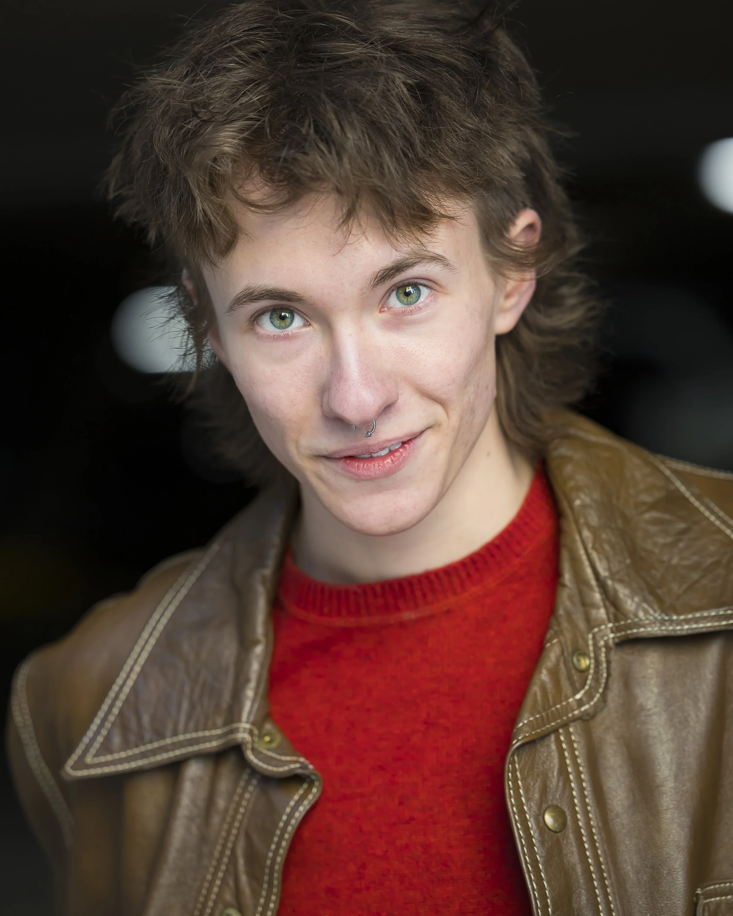 ZEWJ with a short messy mullet wearing a brown jacket and red shirt, looking intensely at camera with a half smirk
