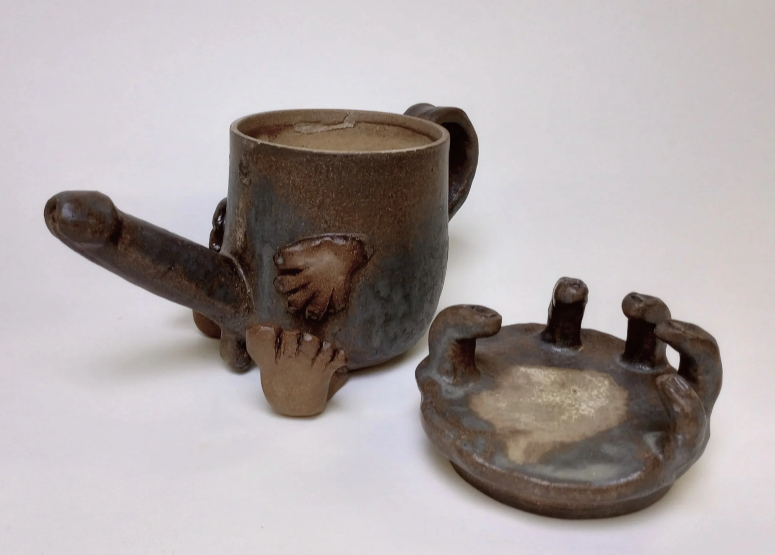 Ceramic coffee pot with arms, legs, and penis spout beside lid with fingers.