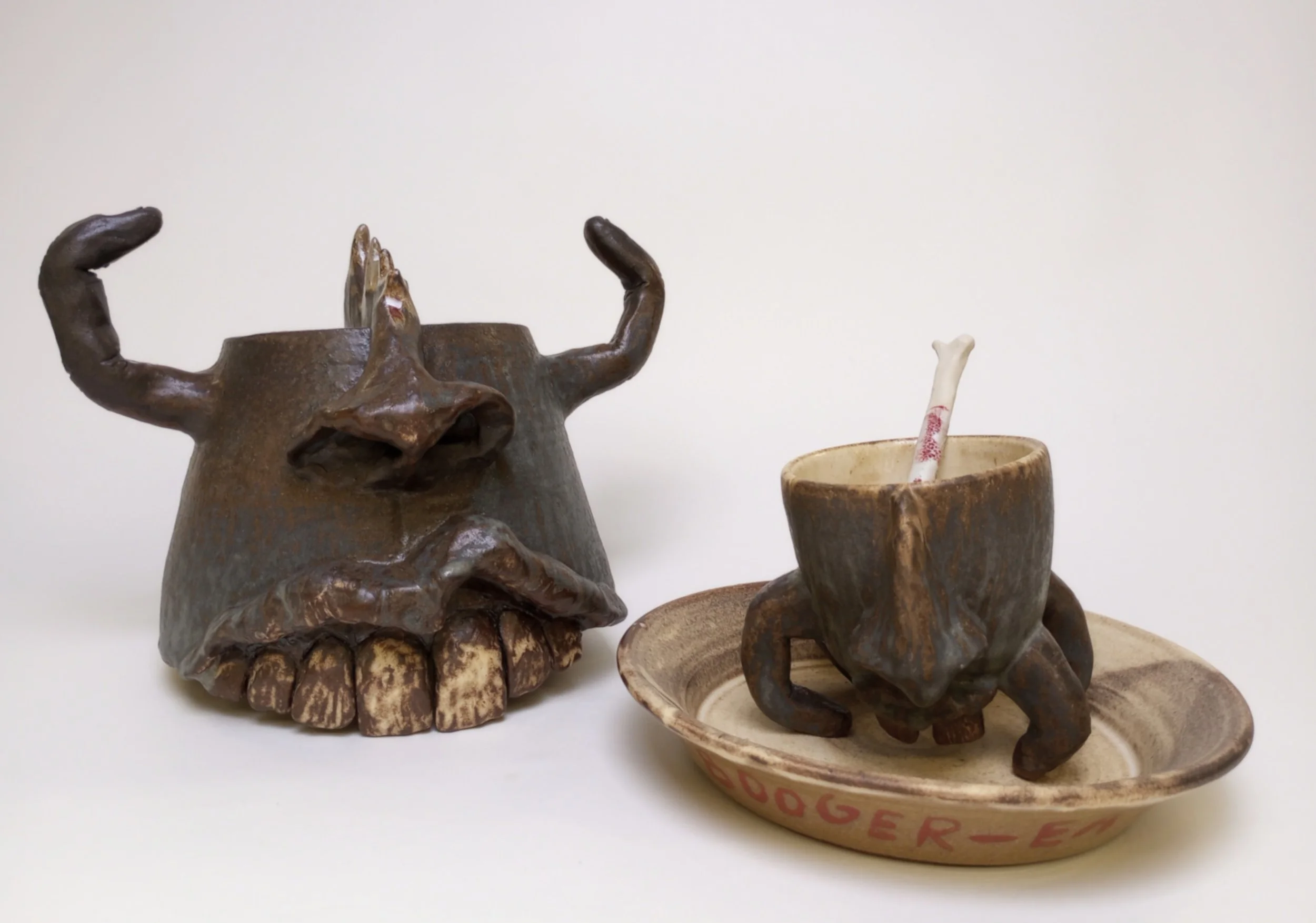 Helmet with big teeth and finger horns beside mug with fingers as legs on a plate.